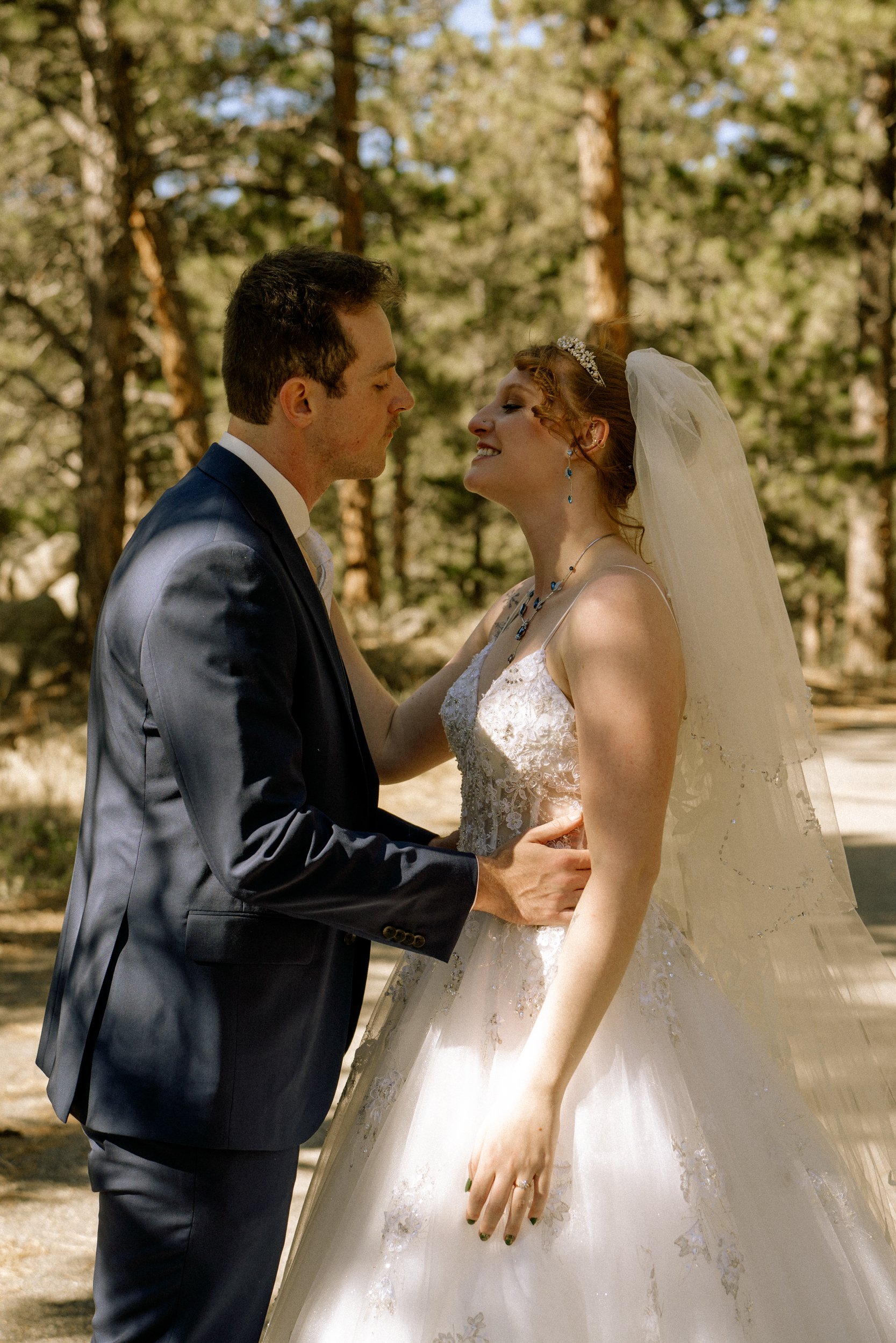 A bride and groom are embracing outdoors in a forest setting, dressed in wedding attire, with trees in the background.