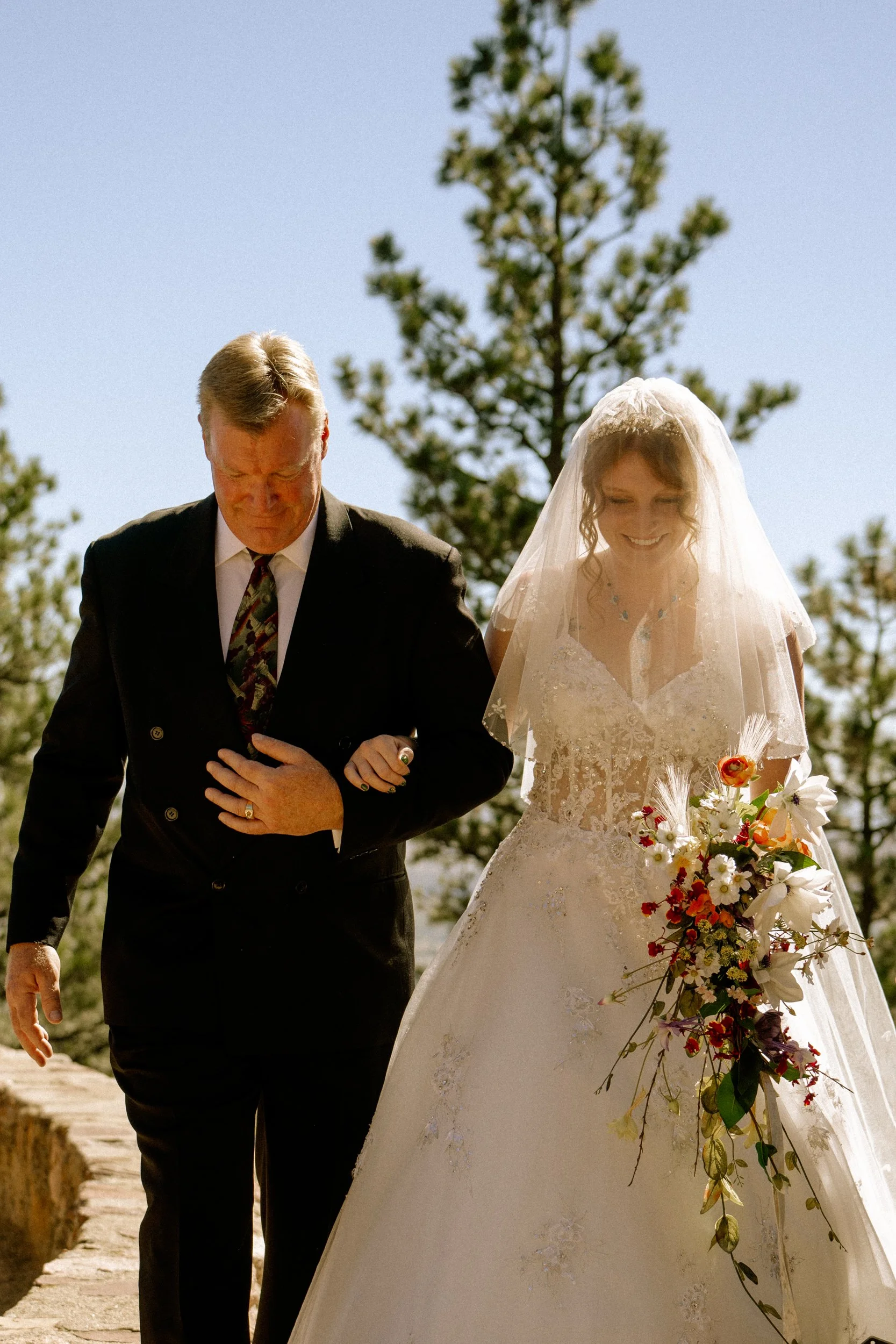 A bride in a wedding gown with lace detail and a veil, holding a bouquet of colorful flowers, walking with an older man in a black suit and colorful tie outdoors on a sunny day, with trees in the background.