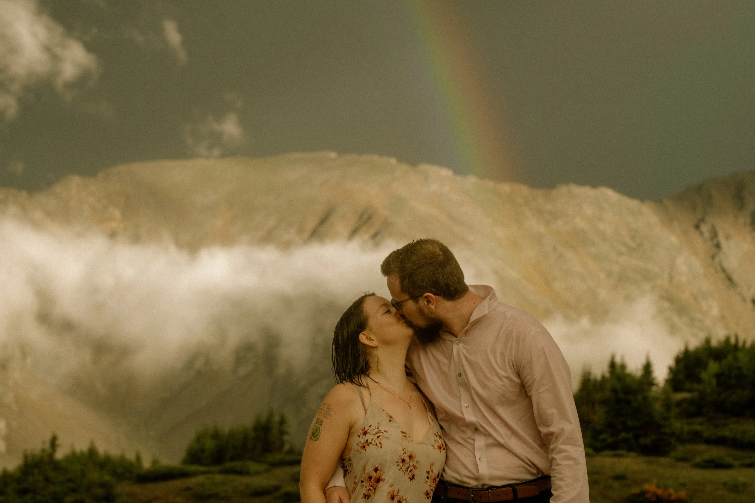 A couple sharing a kiss outdoors with mountains and a rainbow in the background.