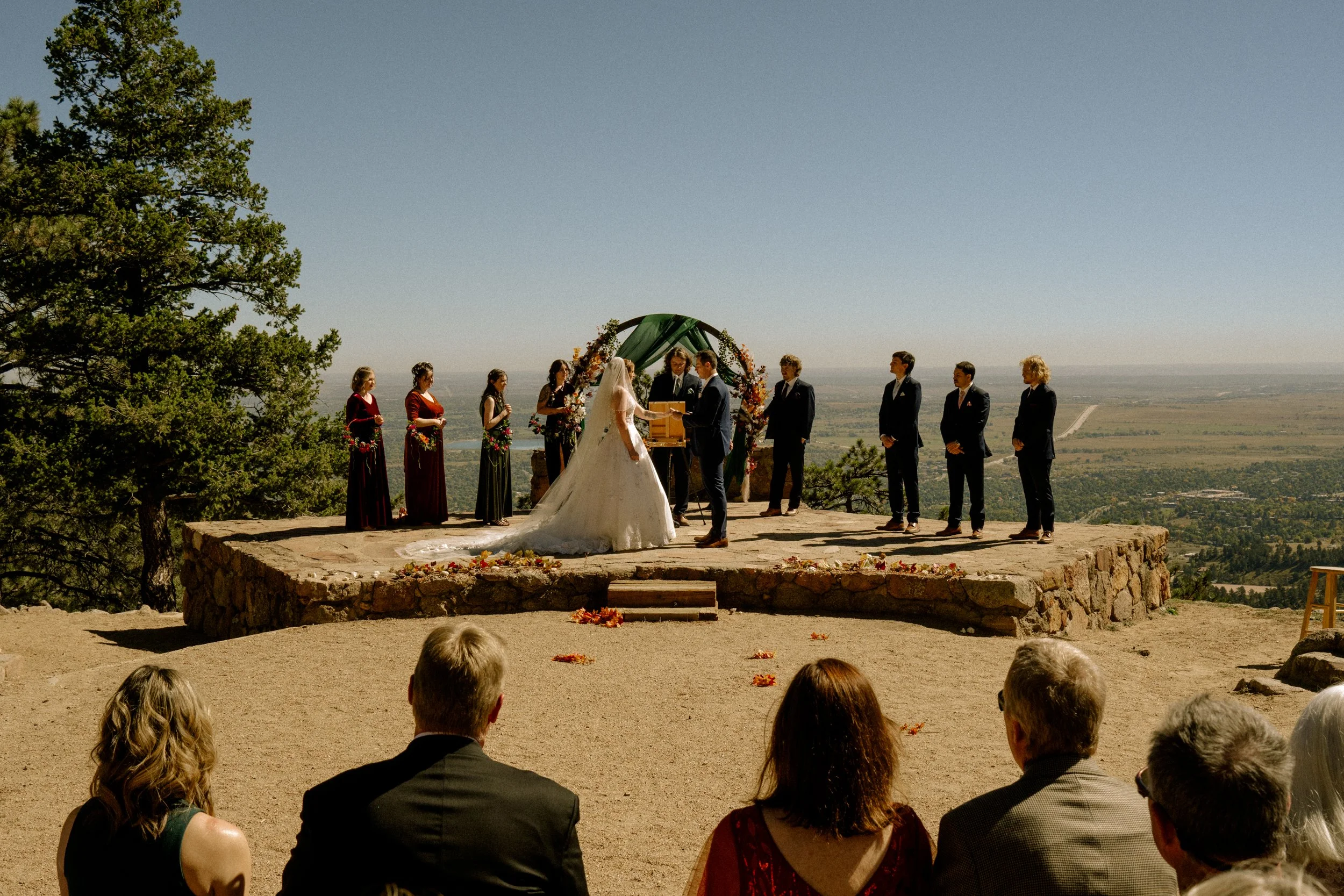 Outdoor wedding ceremony with a bride and groom exchanging vows on a stone platform, surrounded by family and friends, set against a scenic landscape with clear sky and distant fields.