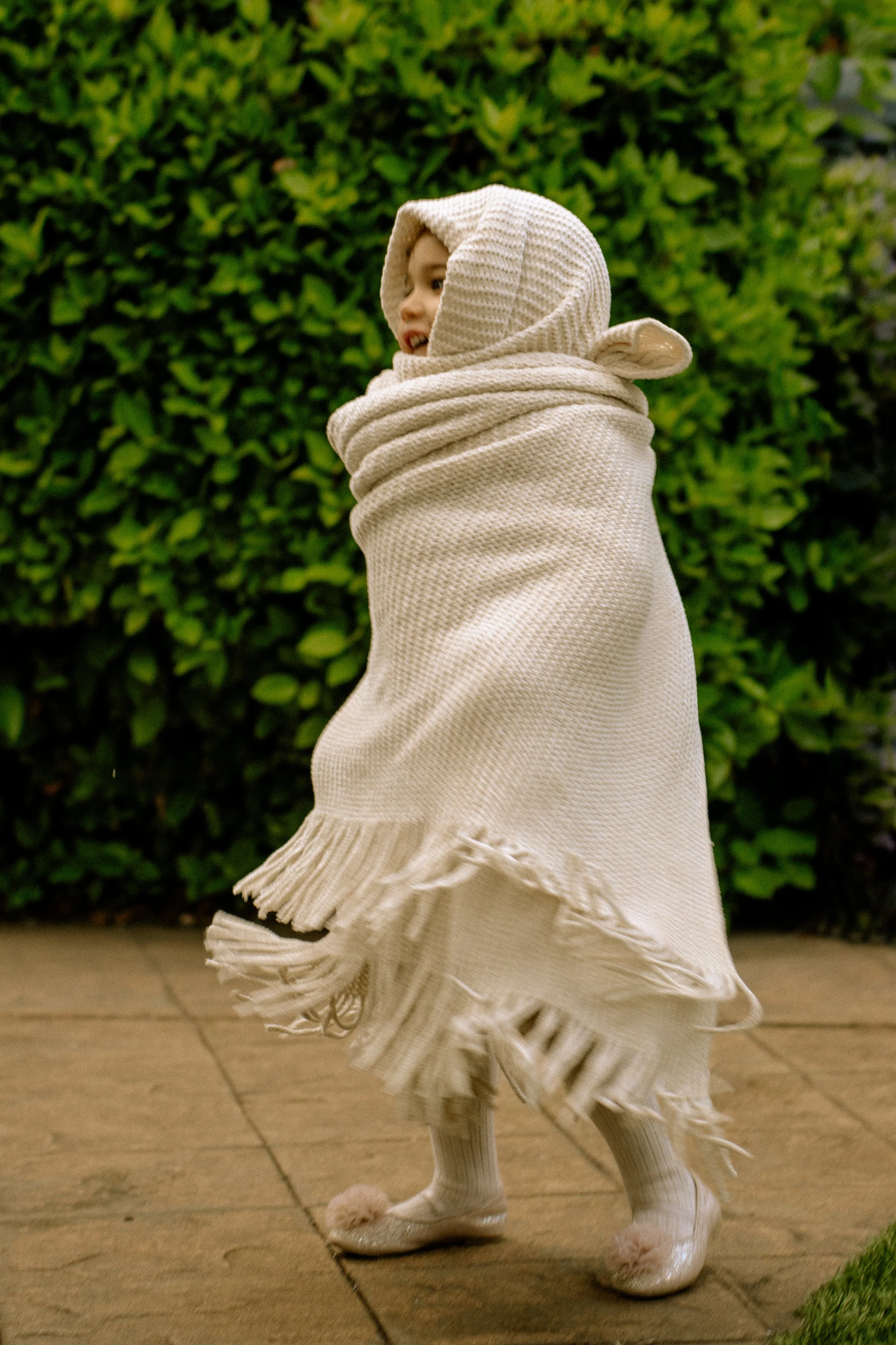 A young girl is wrapped in a cozy white blanket with fringe, wearing a hooded sweater and slippers, standing outdoors near green bushes at night.