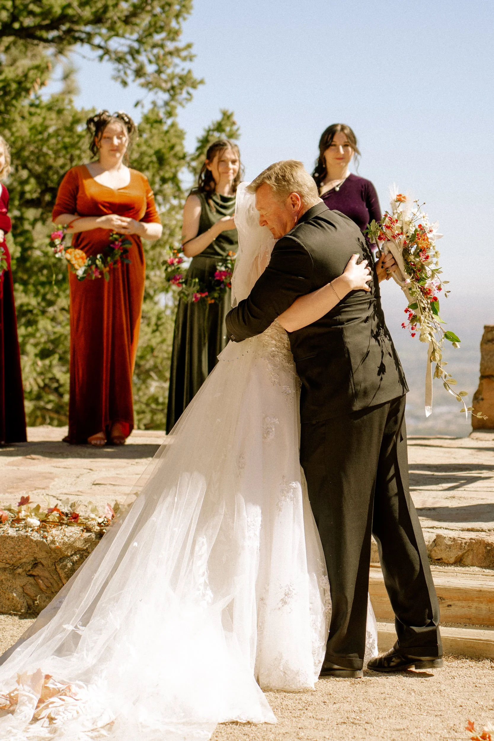 A bride and groom embrace during their outdoor wedding ceremony with bridesmaids in colorful dresses standing behind them.