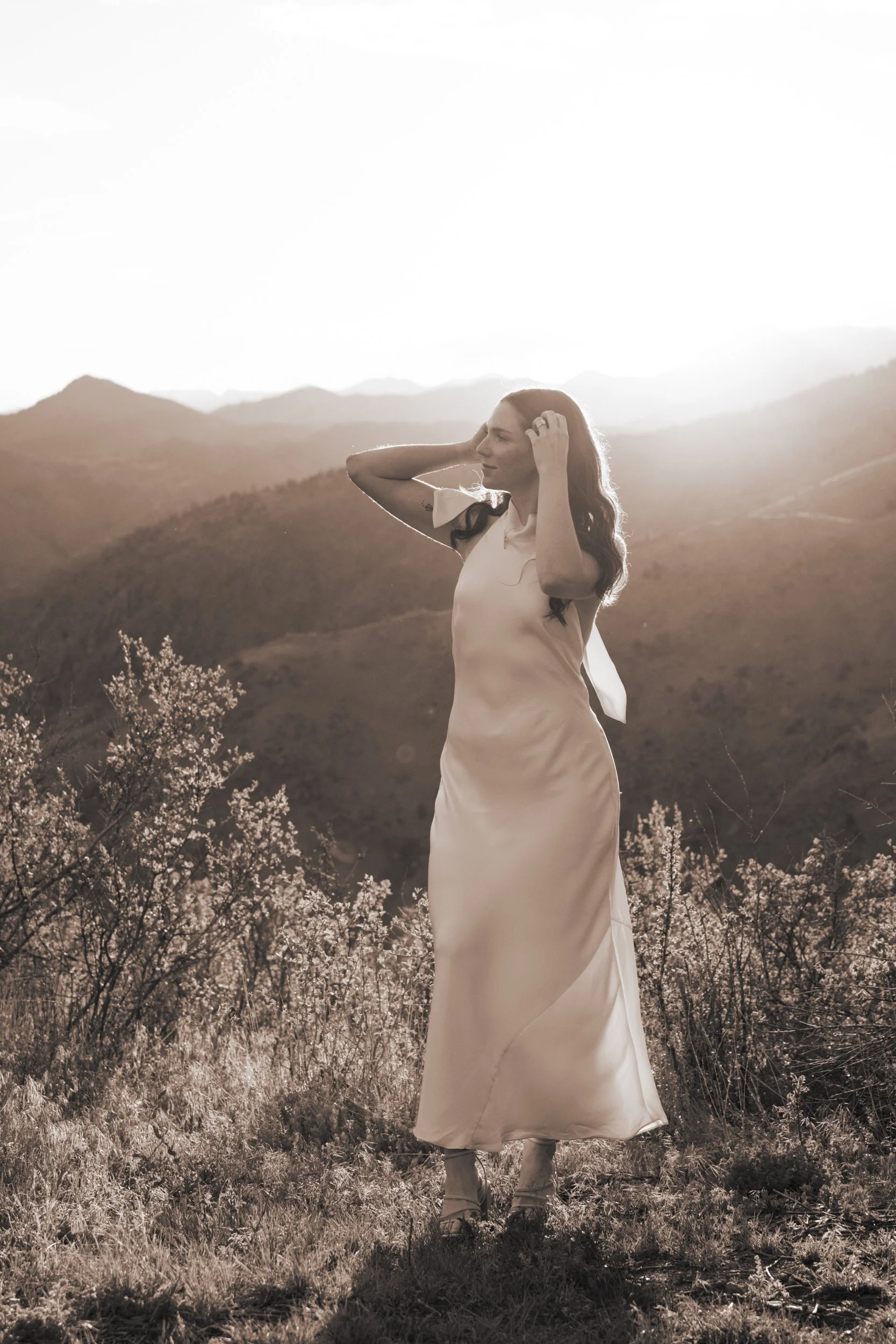 A woman in a white dress standing outdoors on a mountain landscape at sunset, with her hands in her hair and a peaceful expression.