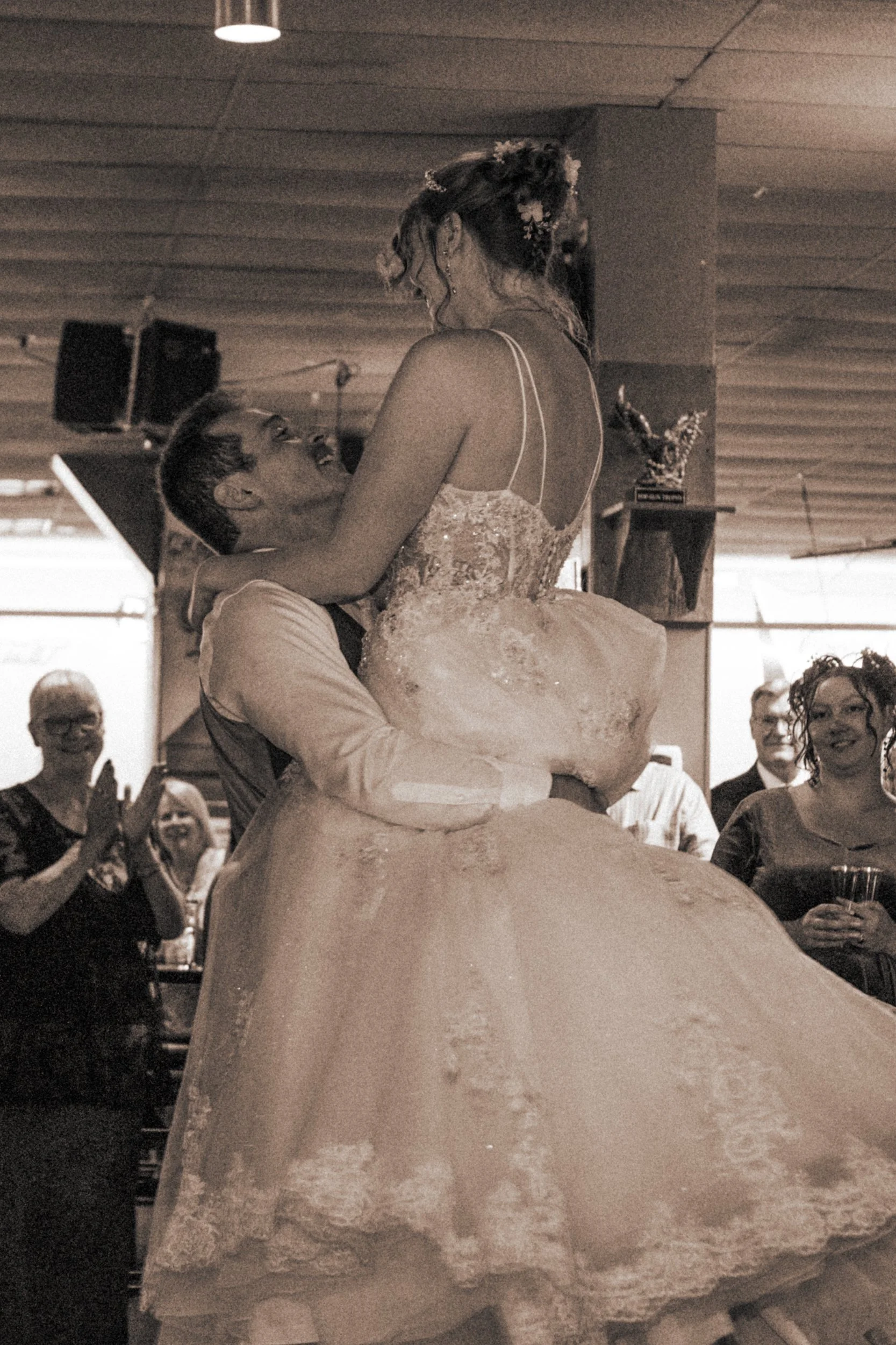 A groom lifts a bride during their wedding reception, surrounded by guests clapping and smiling in a warmly lit room.