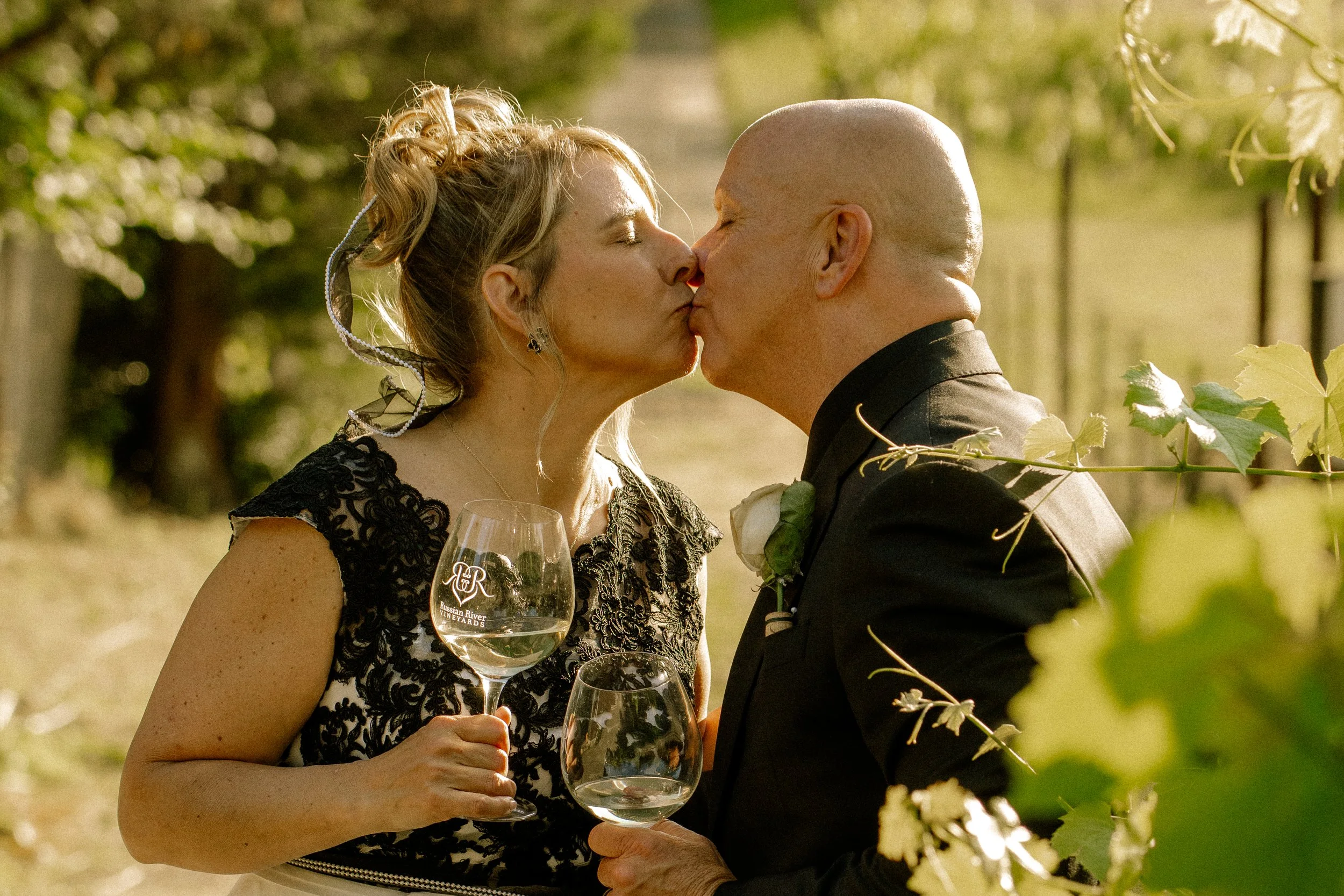 A man and a woman kiss outdoors during daytime, both holding wine glasses with a vineyard logo. The woman has curly blonde hair and wears a black lace dress. The man has a shaved head and wears a black suit with a white rose boutonniere. They are sur