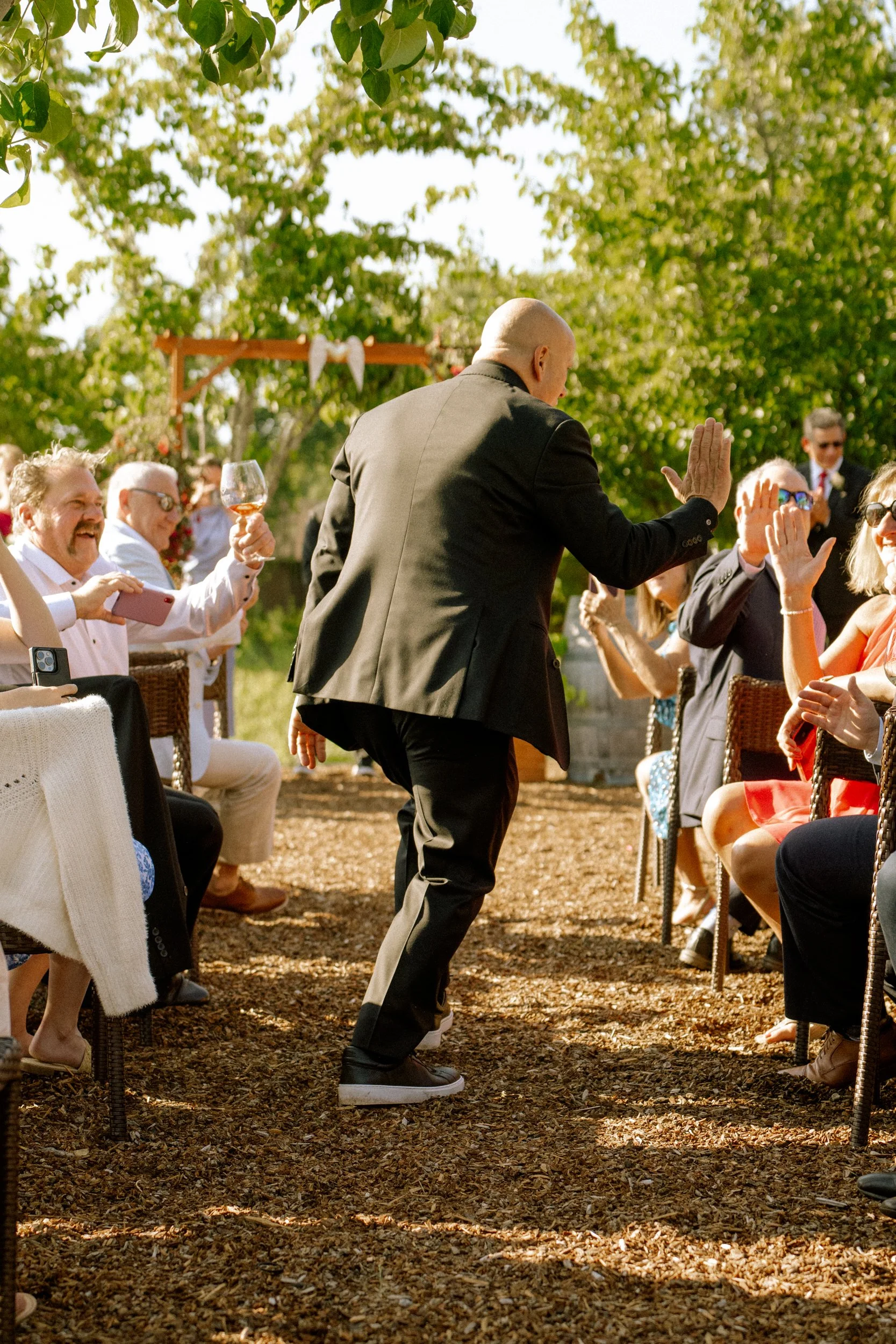 Bald man in black suit and sneakers giving a high five to a woman in a red dress at an outdoor wedding reception surrounded by seated guests under trees.