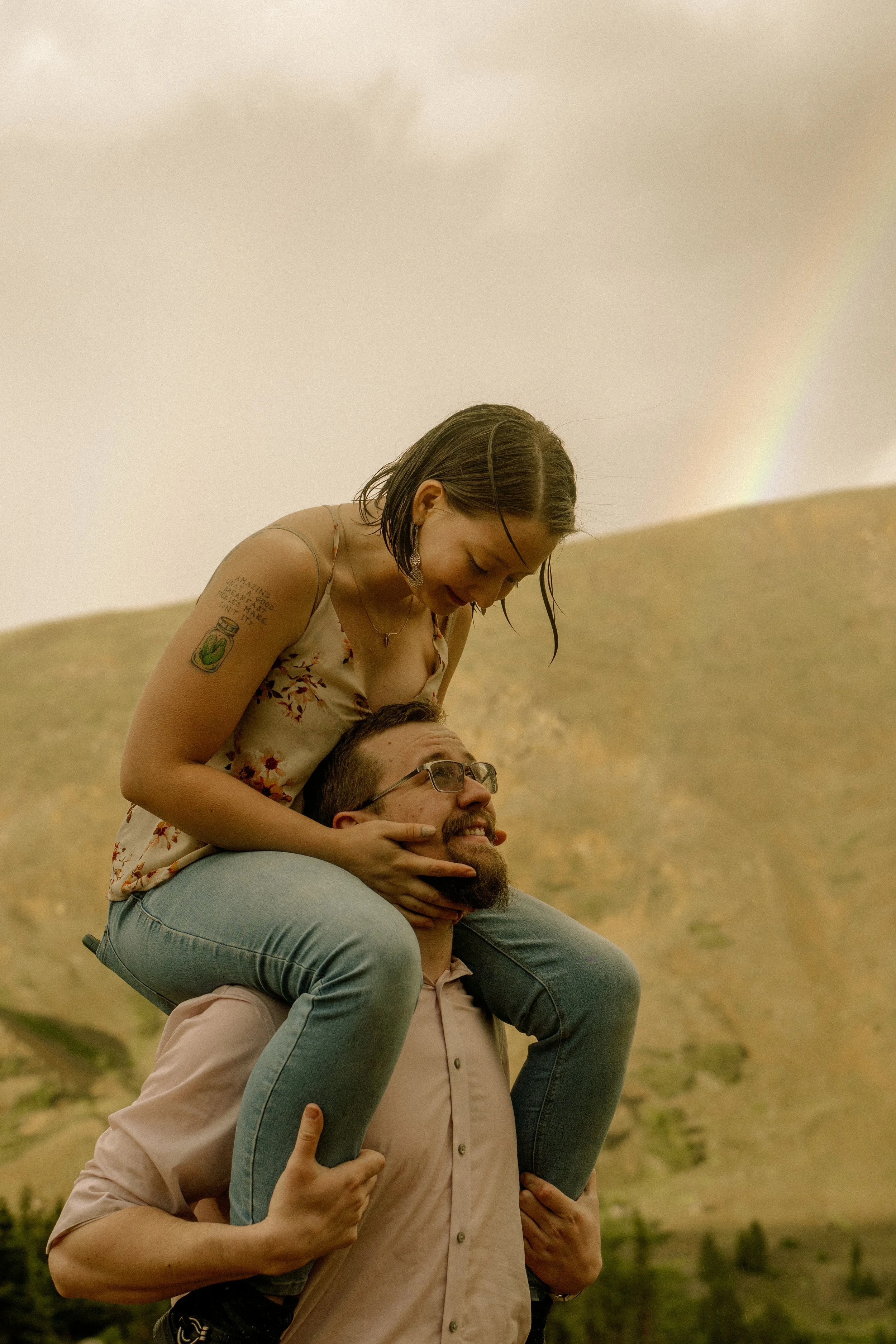 A woman with short dark hair and tattoos on her arm is sitting on a man's shoulders. She is leaning forward and smiling while looking down. The man has glasses, a beard, and is wearing a light-colored shirt. They are outdoors with hills and a rainbow