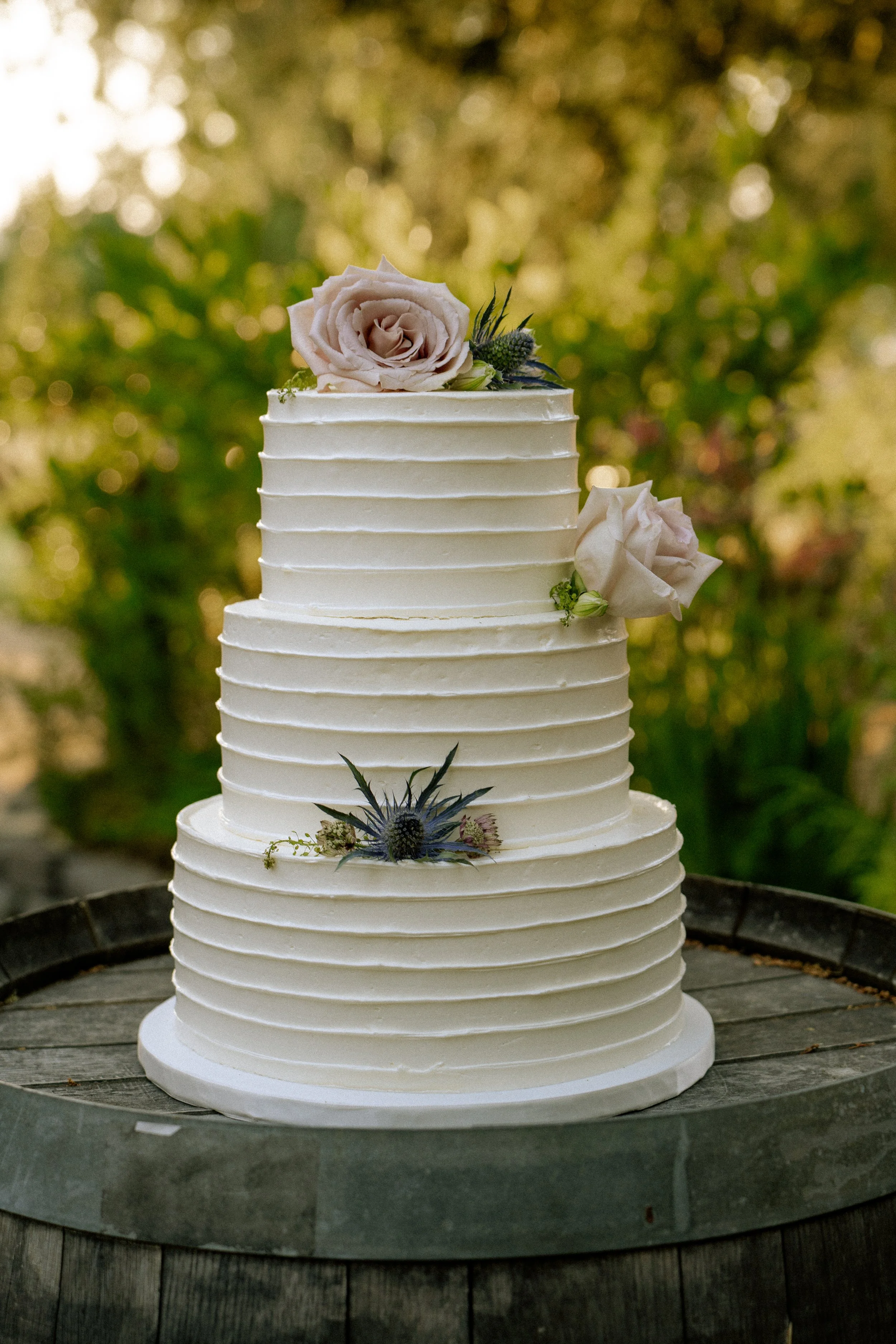 A two-tier white wedding cake decorated with fresh pink and white roses and purple thistle flowers, set outdoors on a wooden surface with green foliage in the background.
