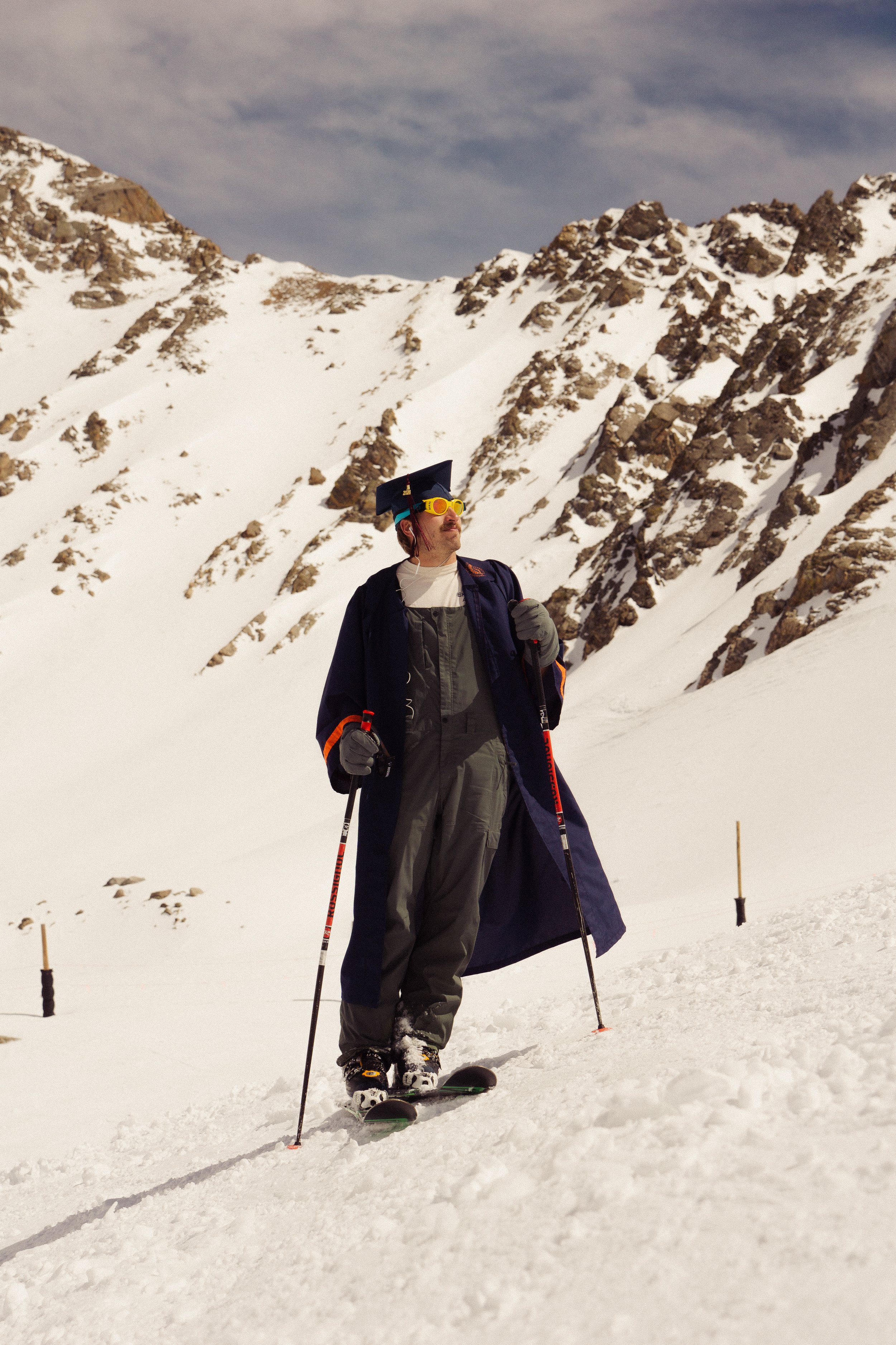 A man in ski gear, including a graduation cap and sunglasses, stands on snow with snow-covered mountains in the background.