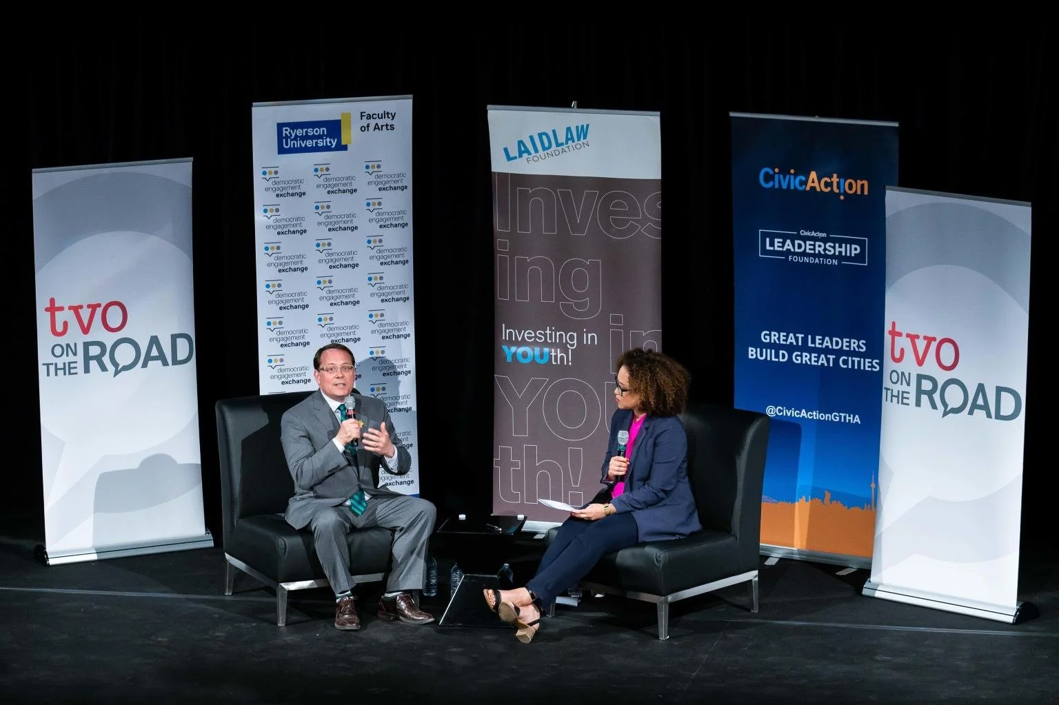 A man and a woman sitting on a stage, engaged in a discussion, with banners behind them displaying logos and slogans related to civic engagement and youth investment.
