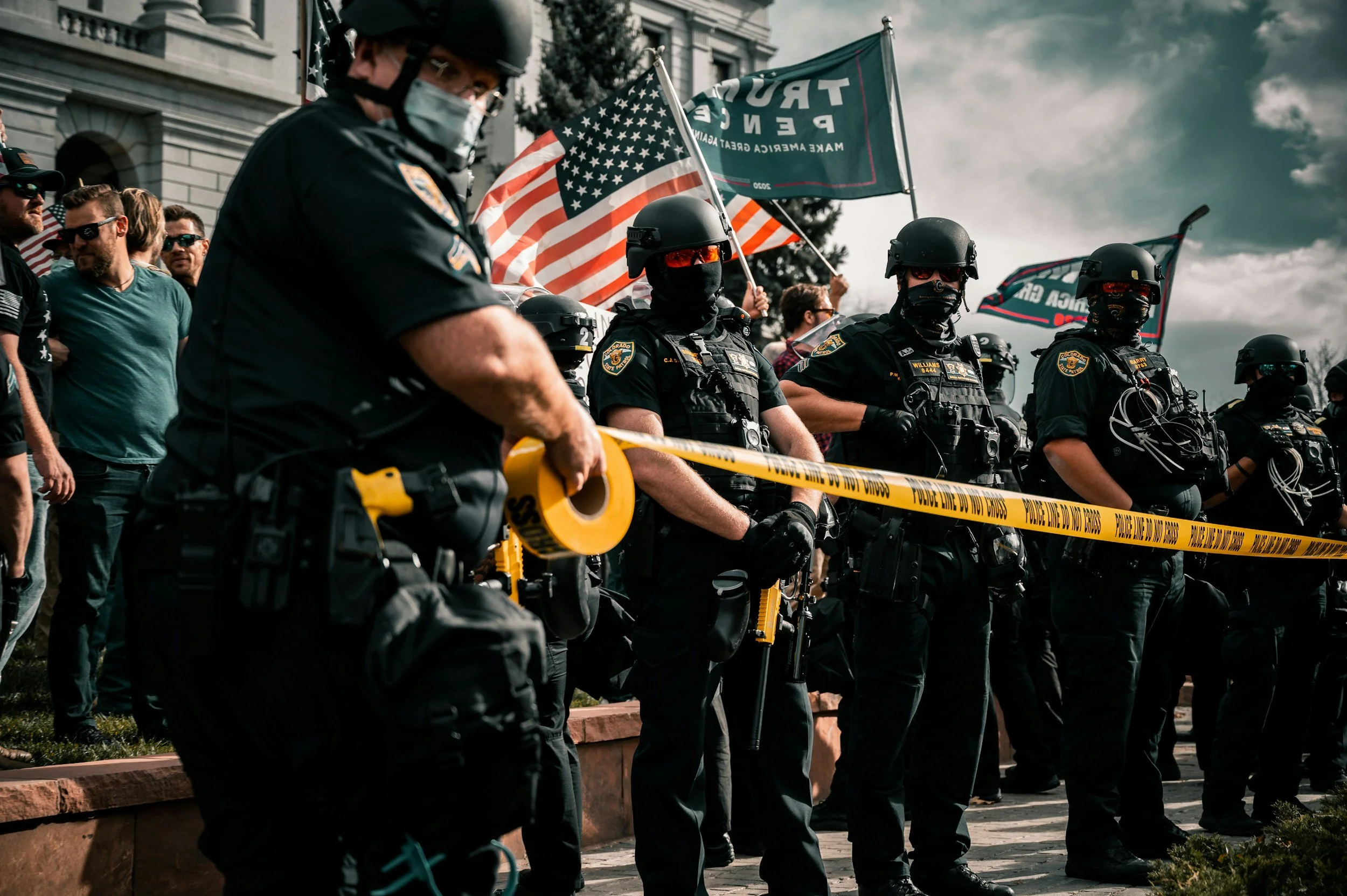 Line of police officers in tactical gear standing behind yellow caution tape, with protesters and flags in the background.