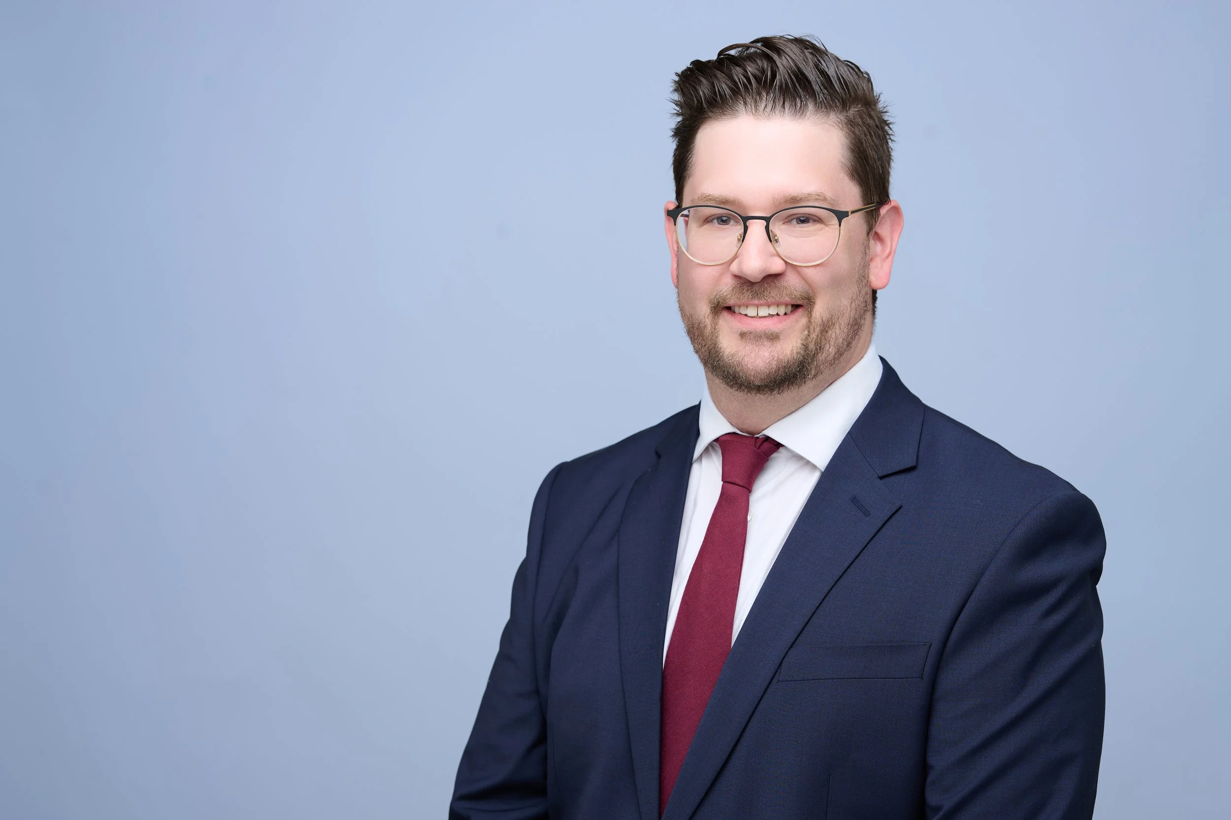 A young man with glasses, short dark hair, a beard, wearing a navy blue suit, white shirt, and maroon tie, smiling against a light blue background.