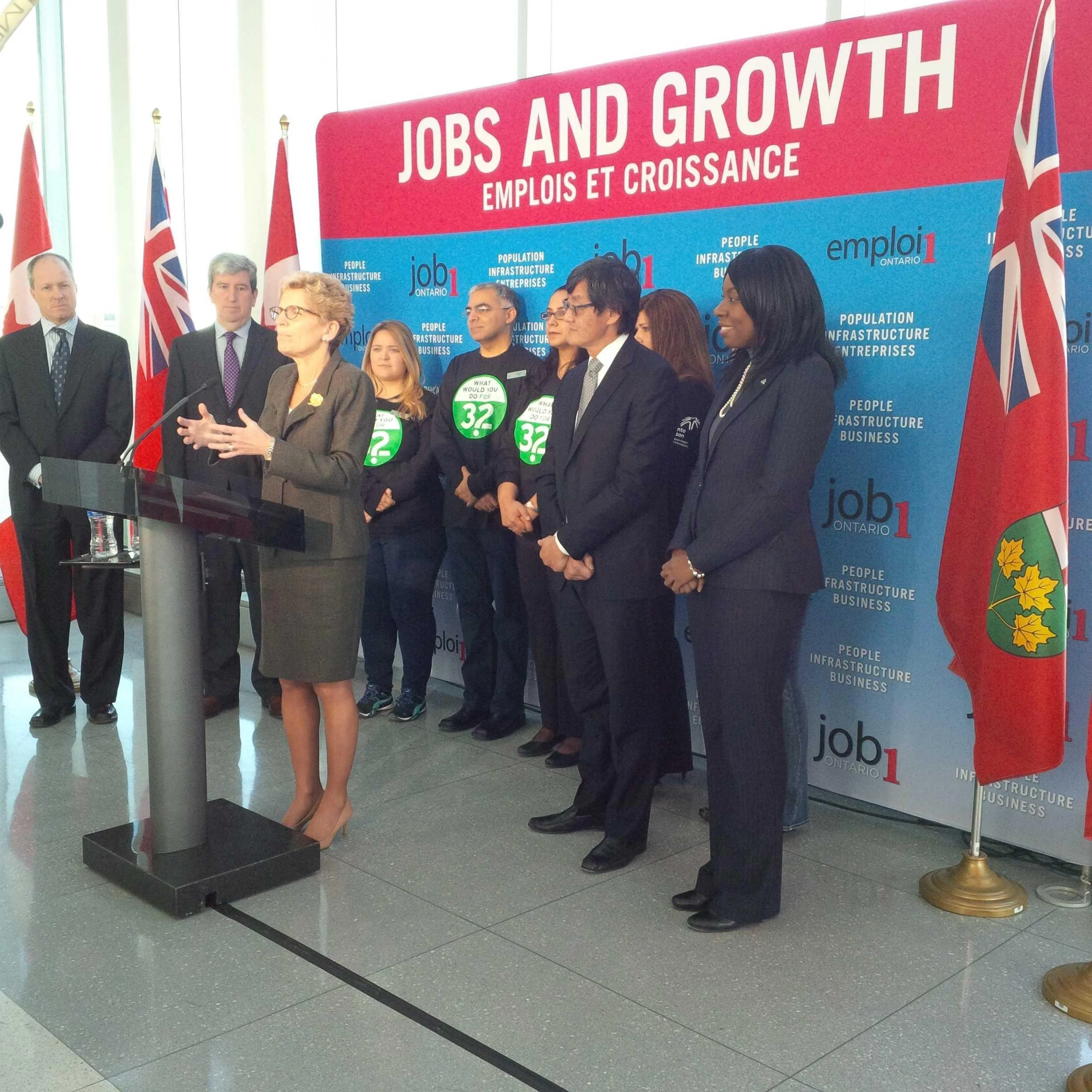 A group of people standing at a press conference with a woman speaking at the podium. The background has a large pink and blue banner with the words "Jobs and Growth" in English and French, and flags are visible. The scene appears to be a formal political or governmental event.