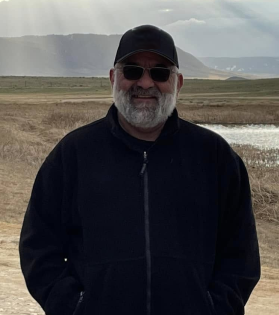 A man with a gray beard wearing sunglasses, a black cap, and a black jacket standing outdoors near a body of water with mountains in the background.