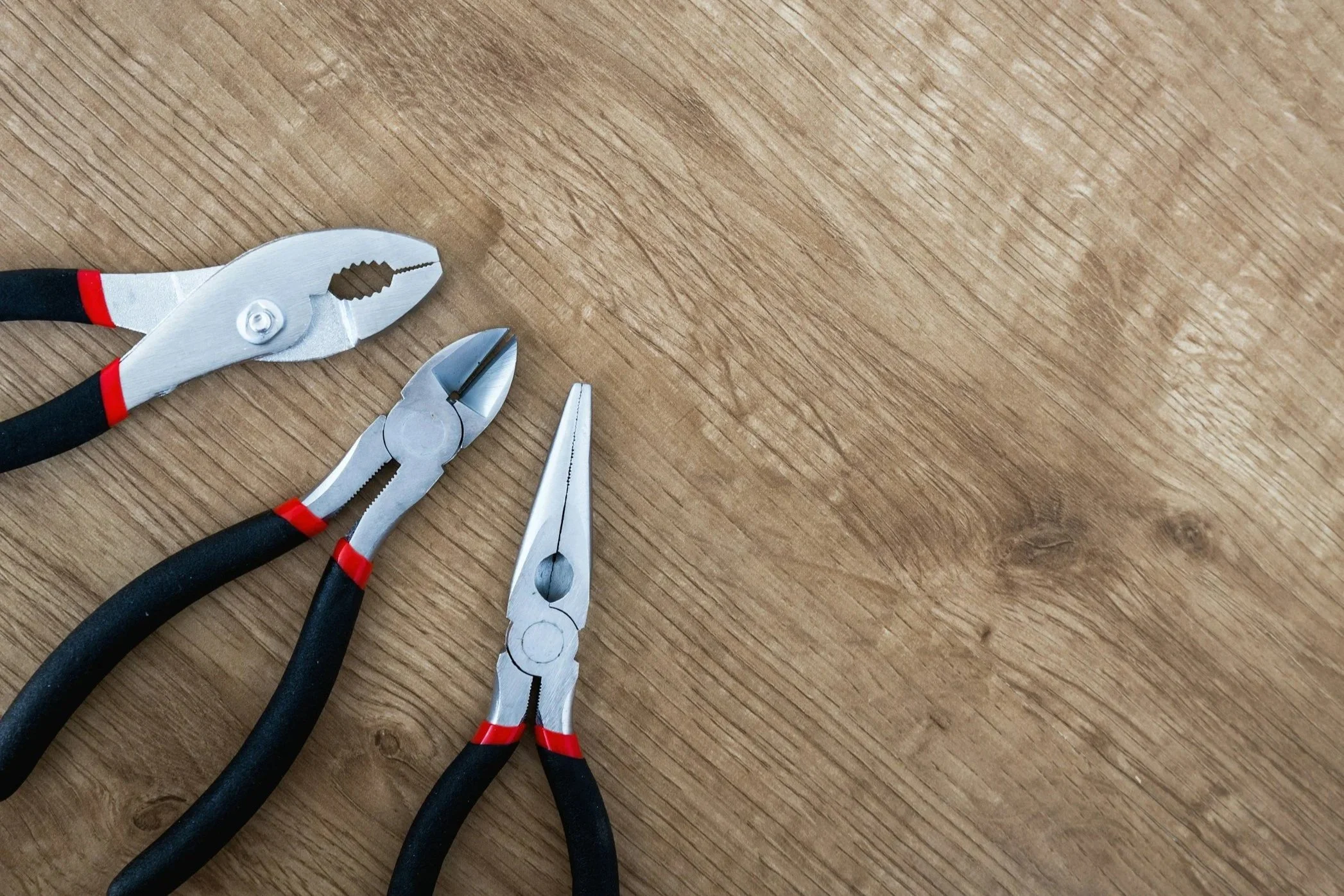 Three needle nose pliers resting on a wooden surface among tools.
