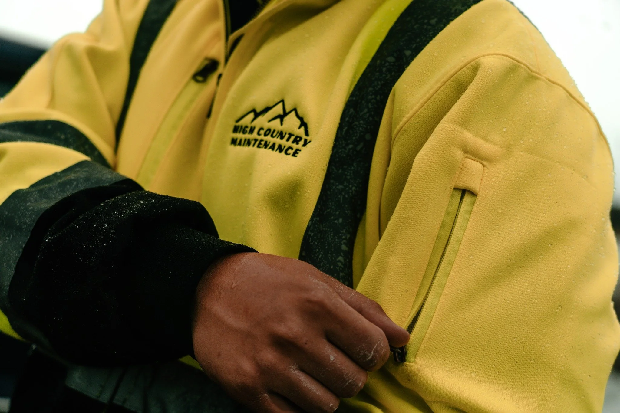 Close-up of a person in a yellow jacket with black accents, labeled 'High Country Maintenance,' adjusting a zipper on the sleeve, with water droplets on the jacket.