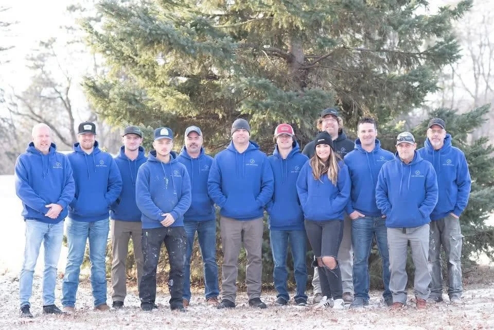 ROC Builders Team members standing outdoors in front of a large pine tree, all wearing matching blue hoodies.