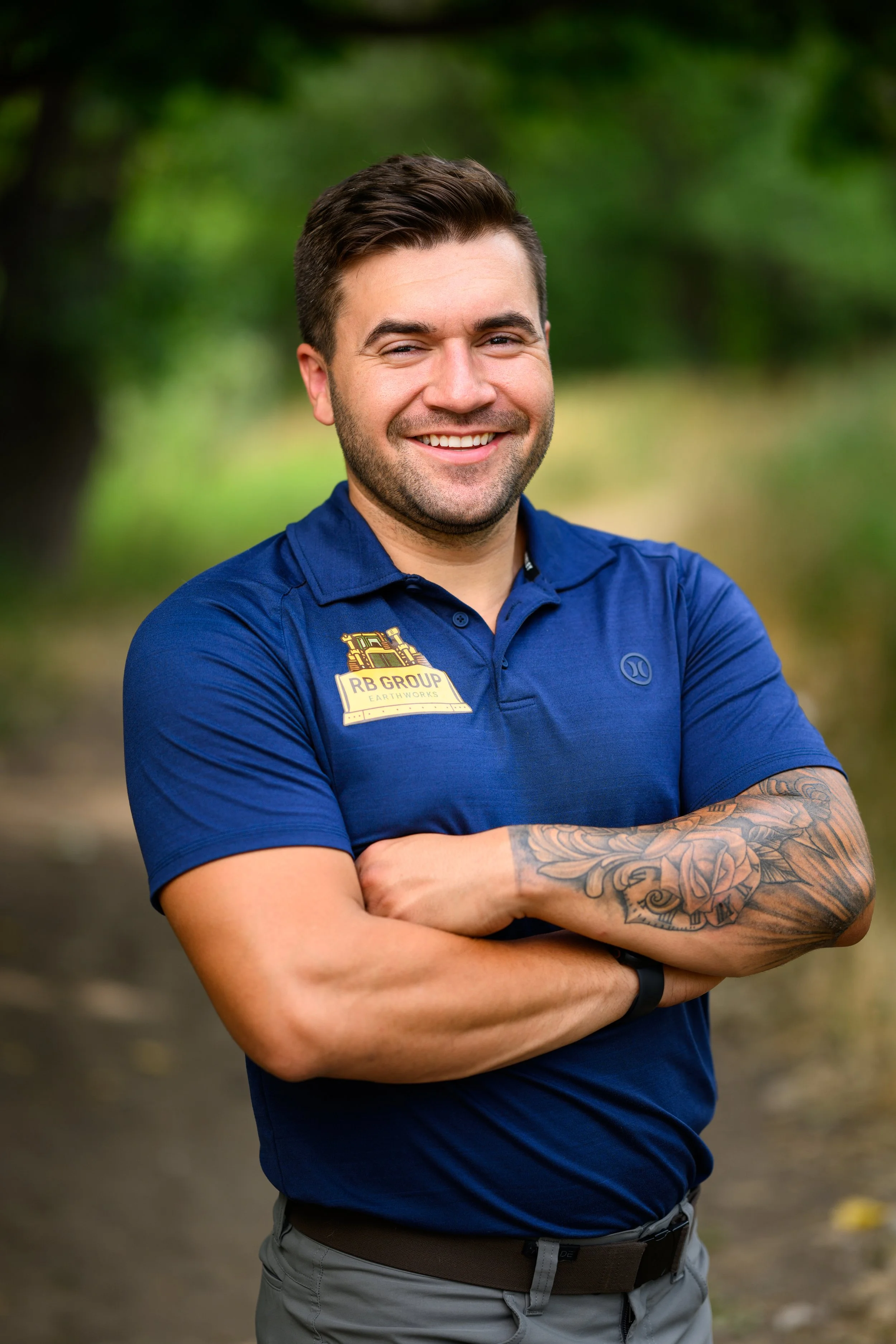 Smiling man with crossed arms, wearing a blue polo shirt with a logo and badge, standing outdoors with blurred green trees in the background.