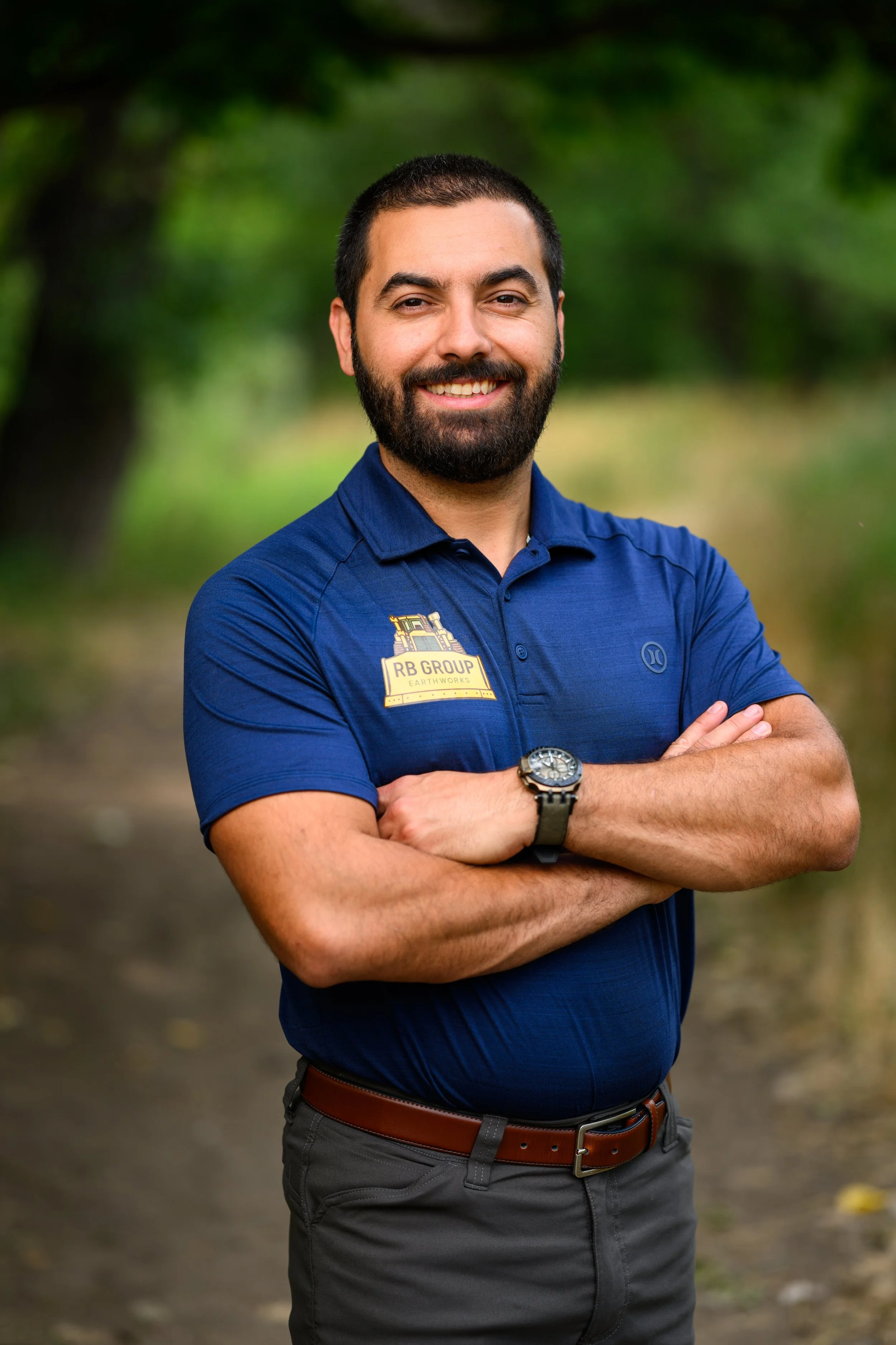 A man with a beard and short hair, smiling, standing outdoors with arms crossed, wearing a blue polo shirt with a logo, gray pants, and a watch, in a natural setting with green trees in the background.