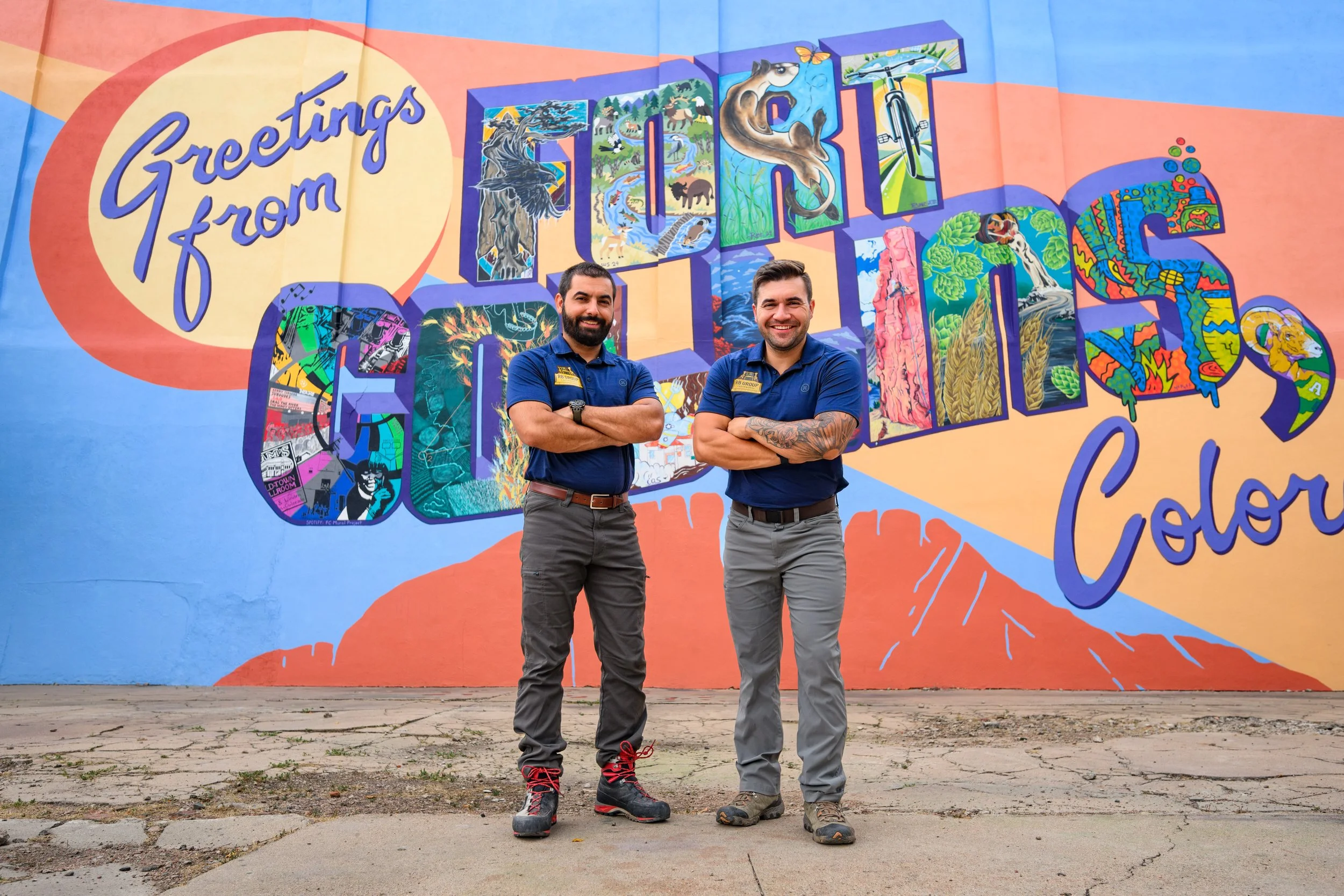 Two men standing in front of a colorful mural that says "Greetings from Tacoma, Washington" and features various illustrated landmarks and scenes.