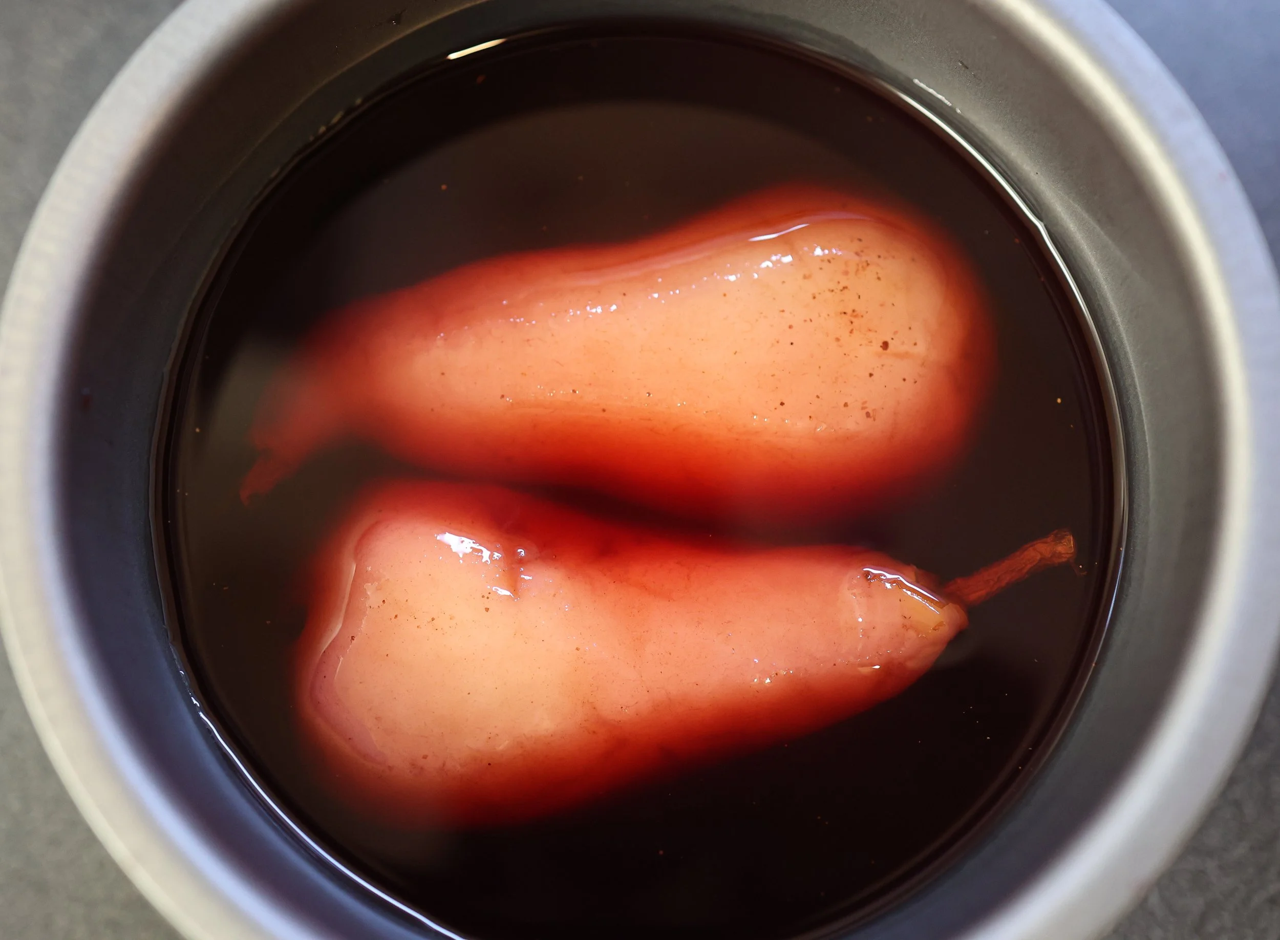 A top-down view of two peeled pears soaking in dark red liquid in a metal container.