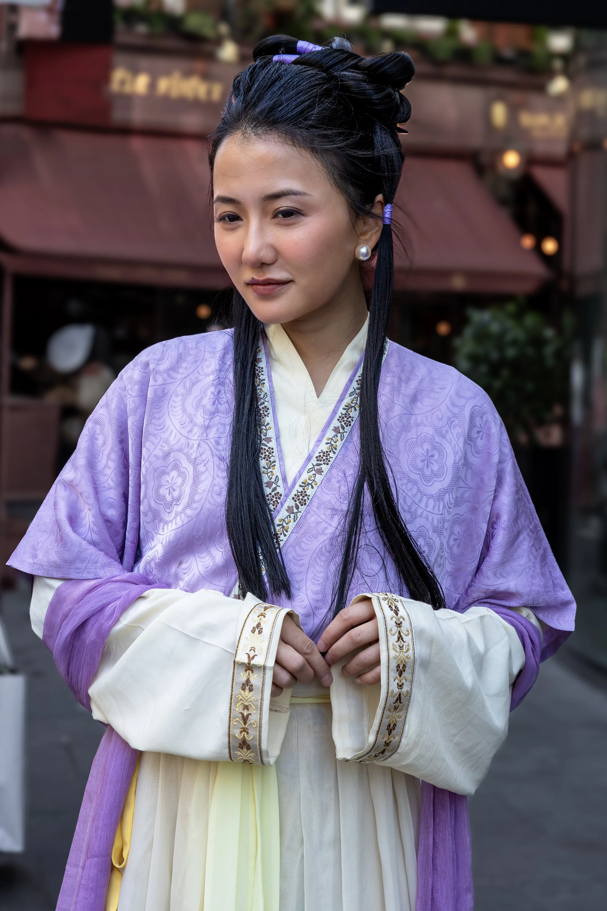 Woman wearing traditional Asian attire with a purple top, cream-colored inner garment, and long black hair in an elaborate style, standing in an outdoor market.