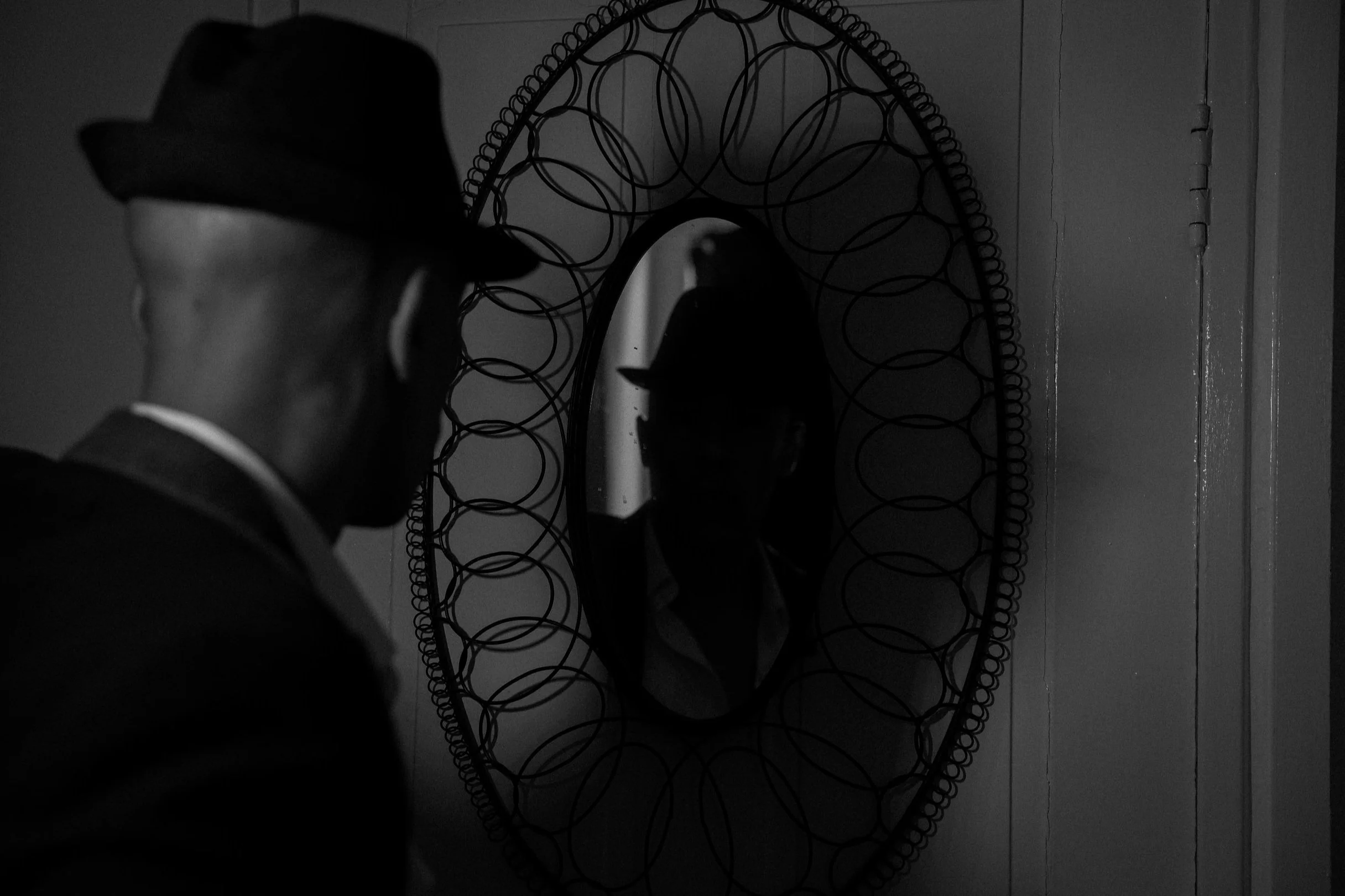 A man wearing a hat is looking at his reflection in an oval mirror with a decorative surrounding frame, mounted on a wall.