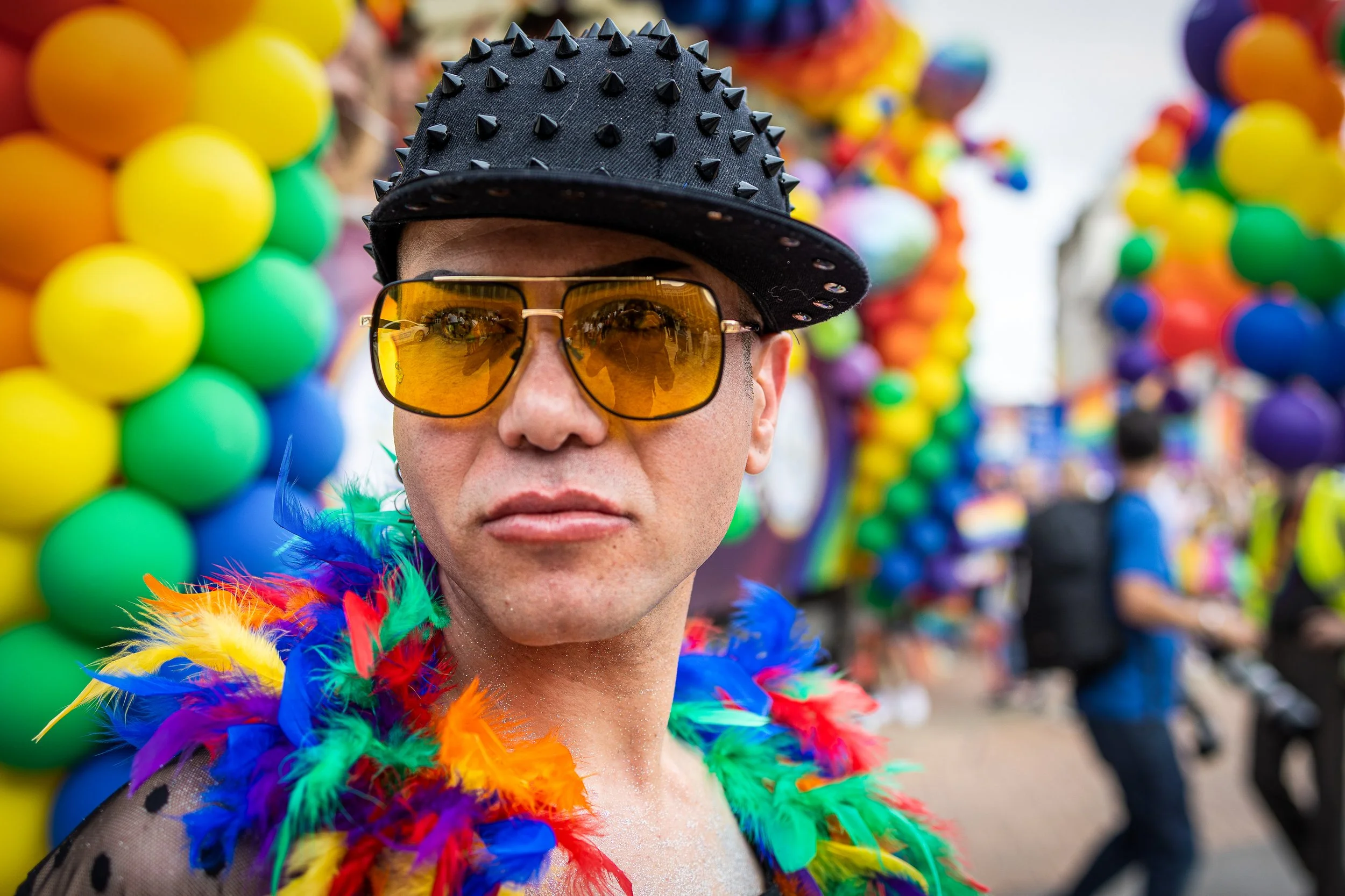 Person wearing yellow sunglasses, a black spiked cap, and a rainbow feather boa at a pride parade with rainbow balloons in the background.
