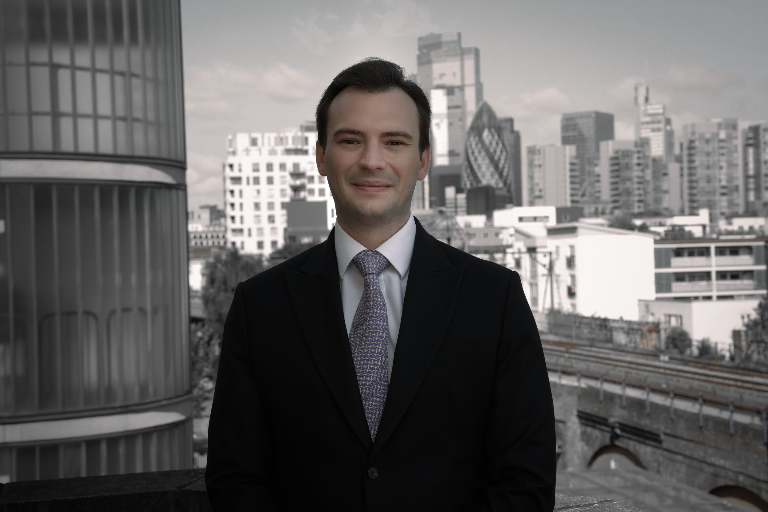 A man in a suit smiling, standing outdoors with a city skyline in the background.