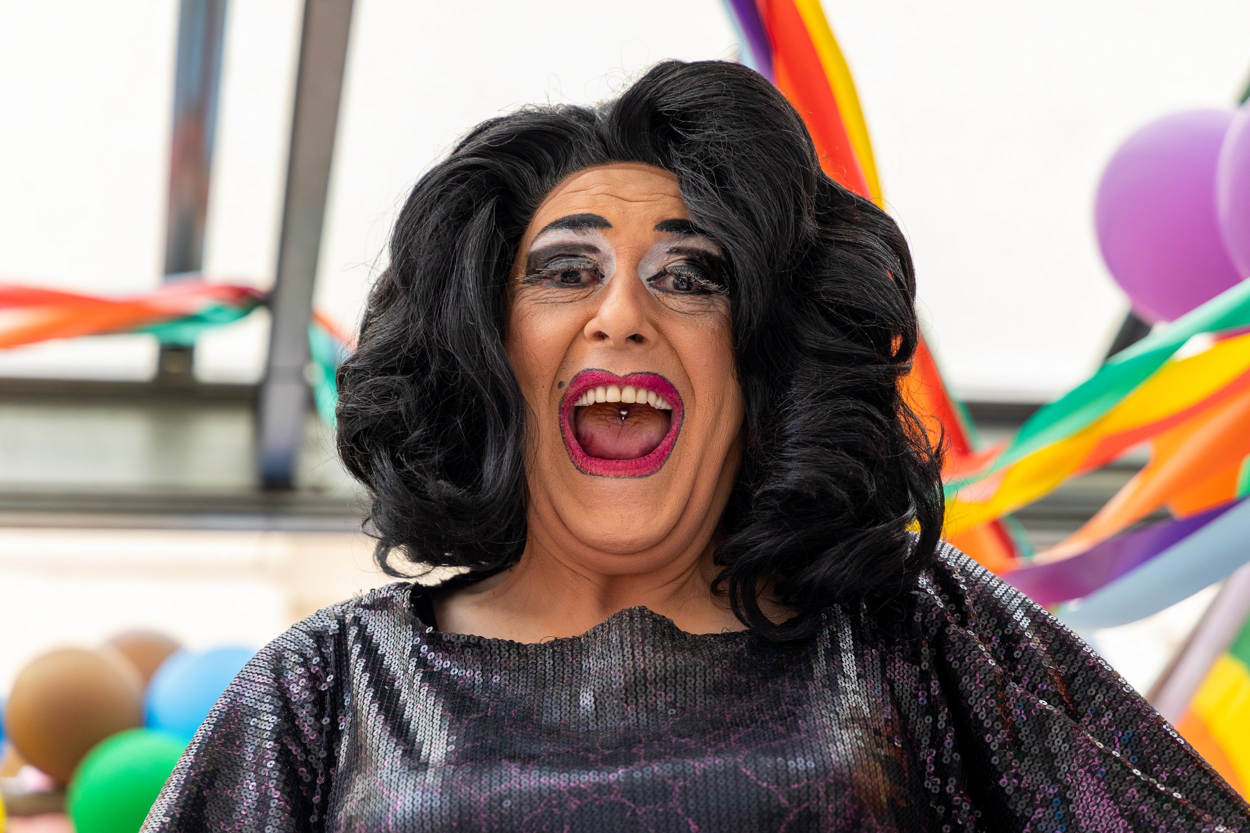 A person with long black hair, makeup, and lip color, dressed in a shiny, dark outfit, with a colorful carnival or fair background with balloons and streamers.