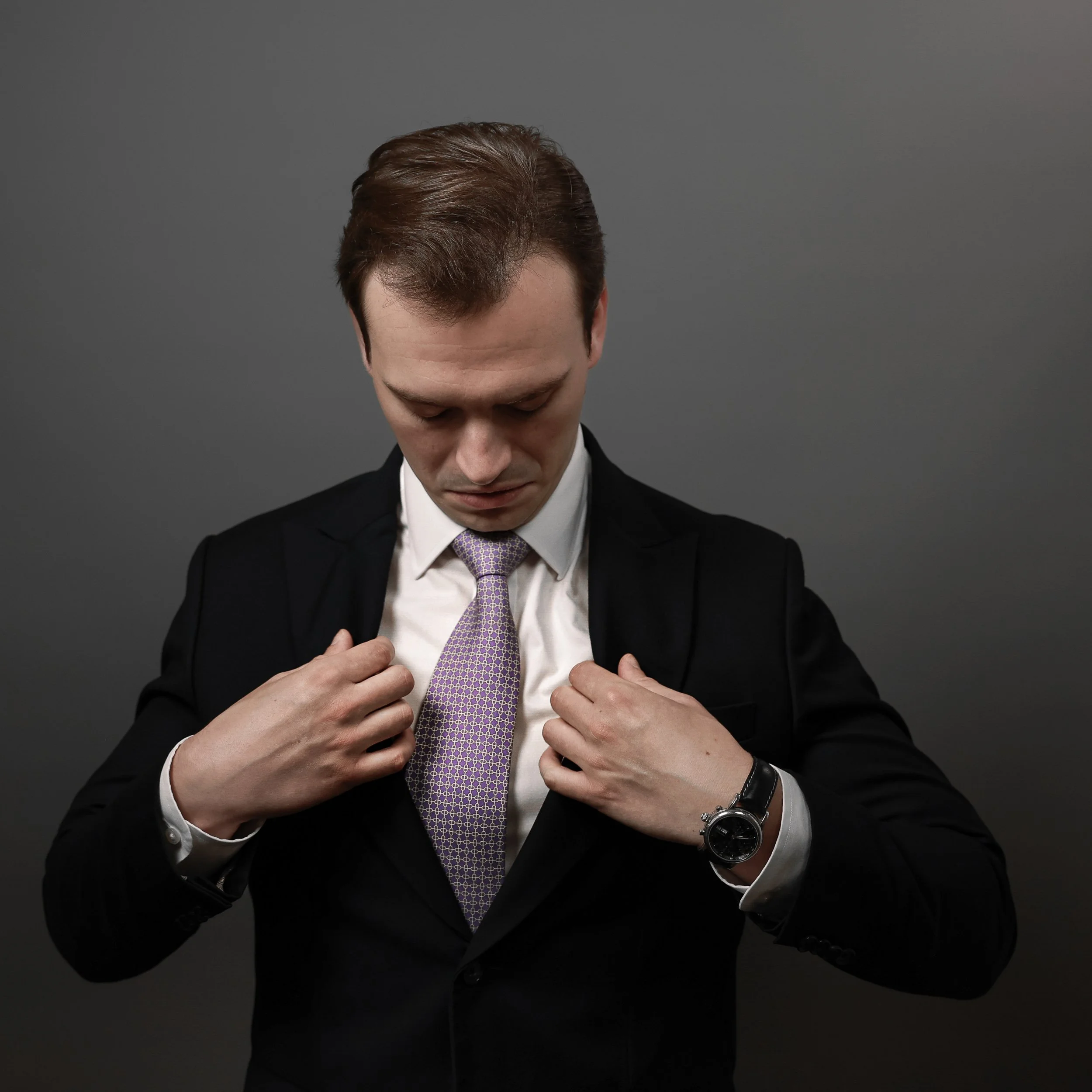 Man in black suit adjusting his purple patterned tie in front of gray background