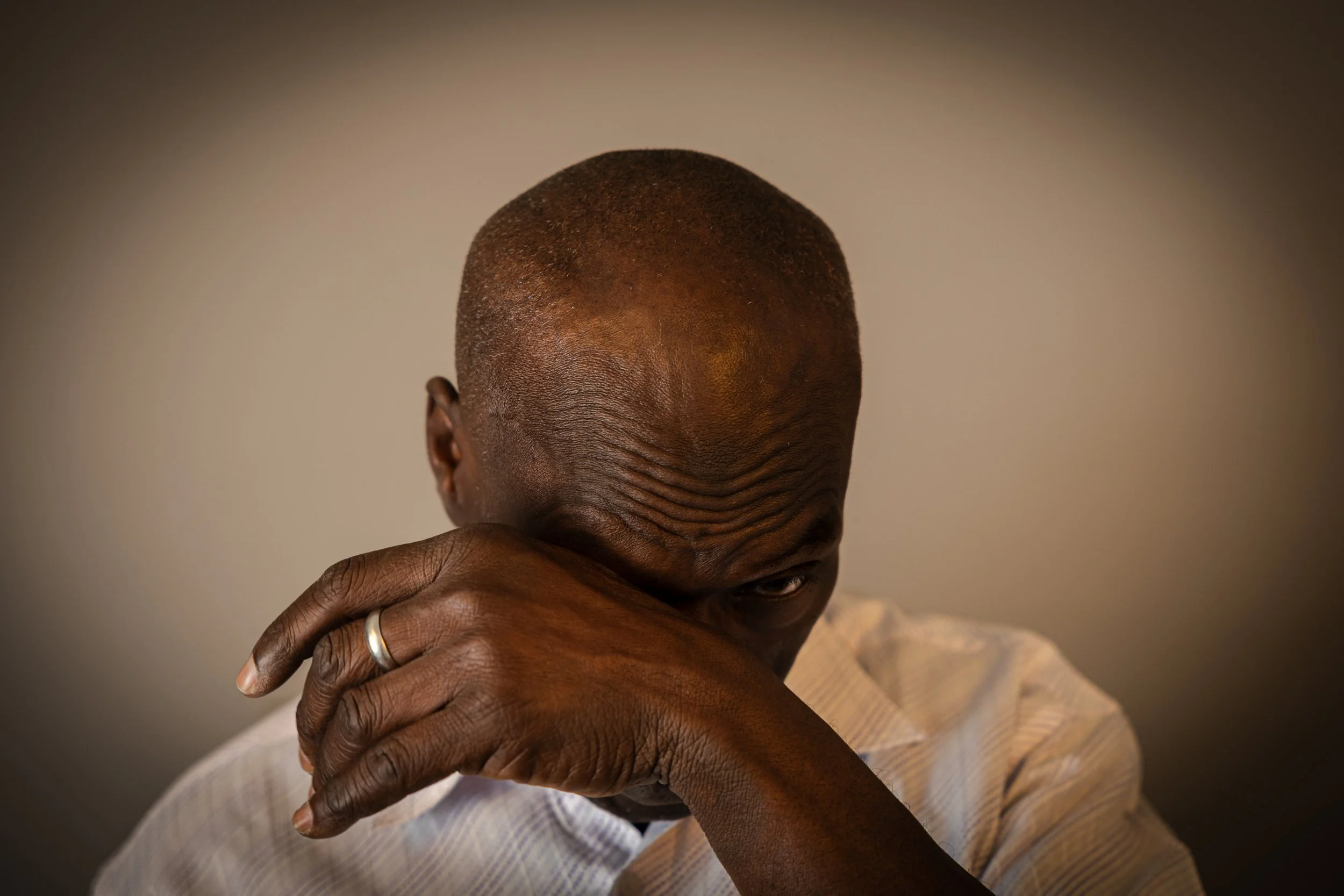 A man with a shaved head resting his forehead on his hand, which has a wedding ring, against a plain background.