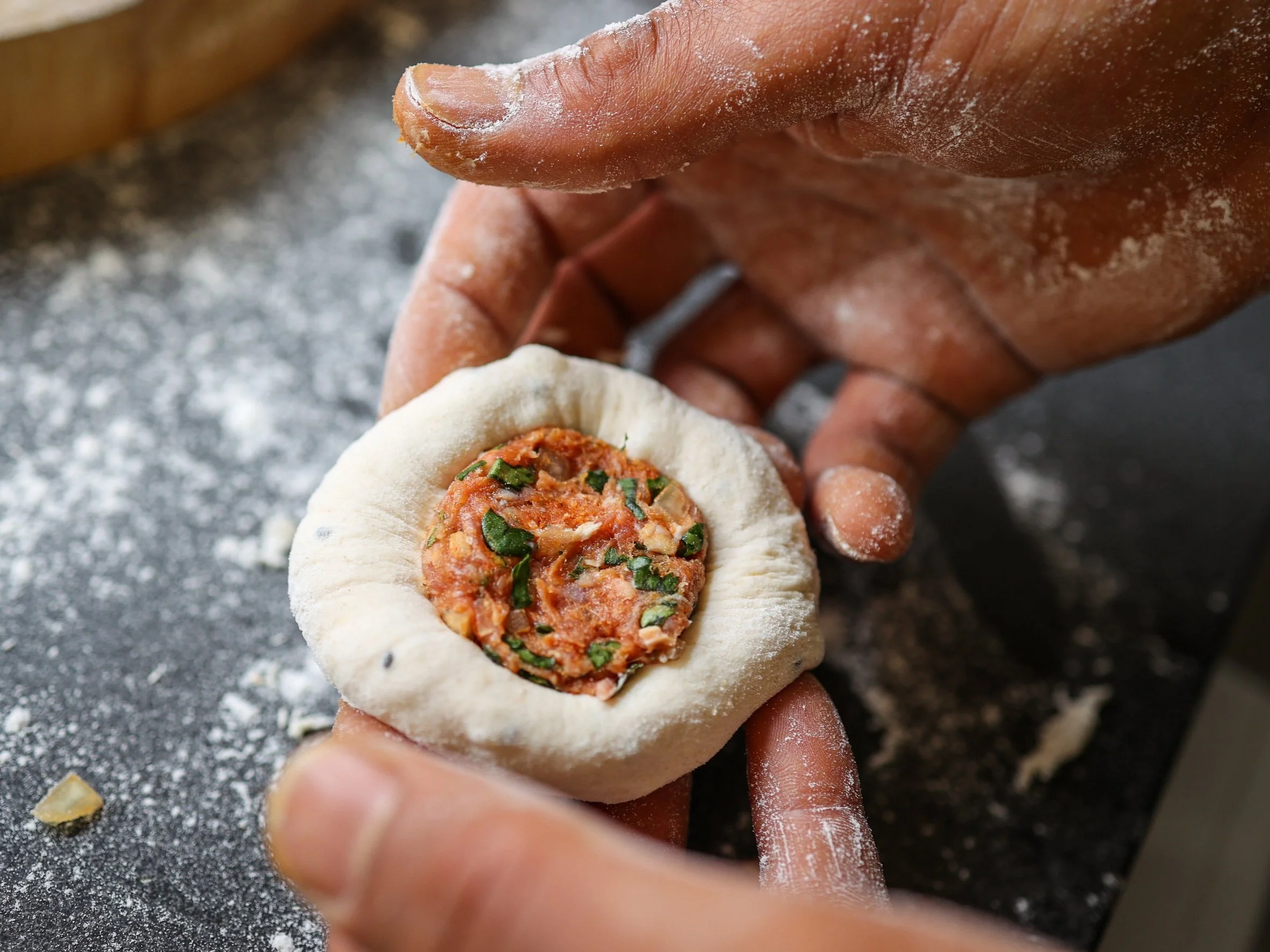 Hands shaping a piece of dough around a filling of tomato sauce, herbs, and other ingredients on a floured surface.