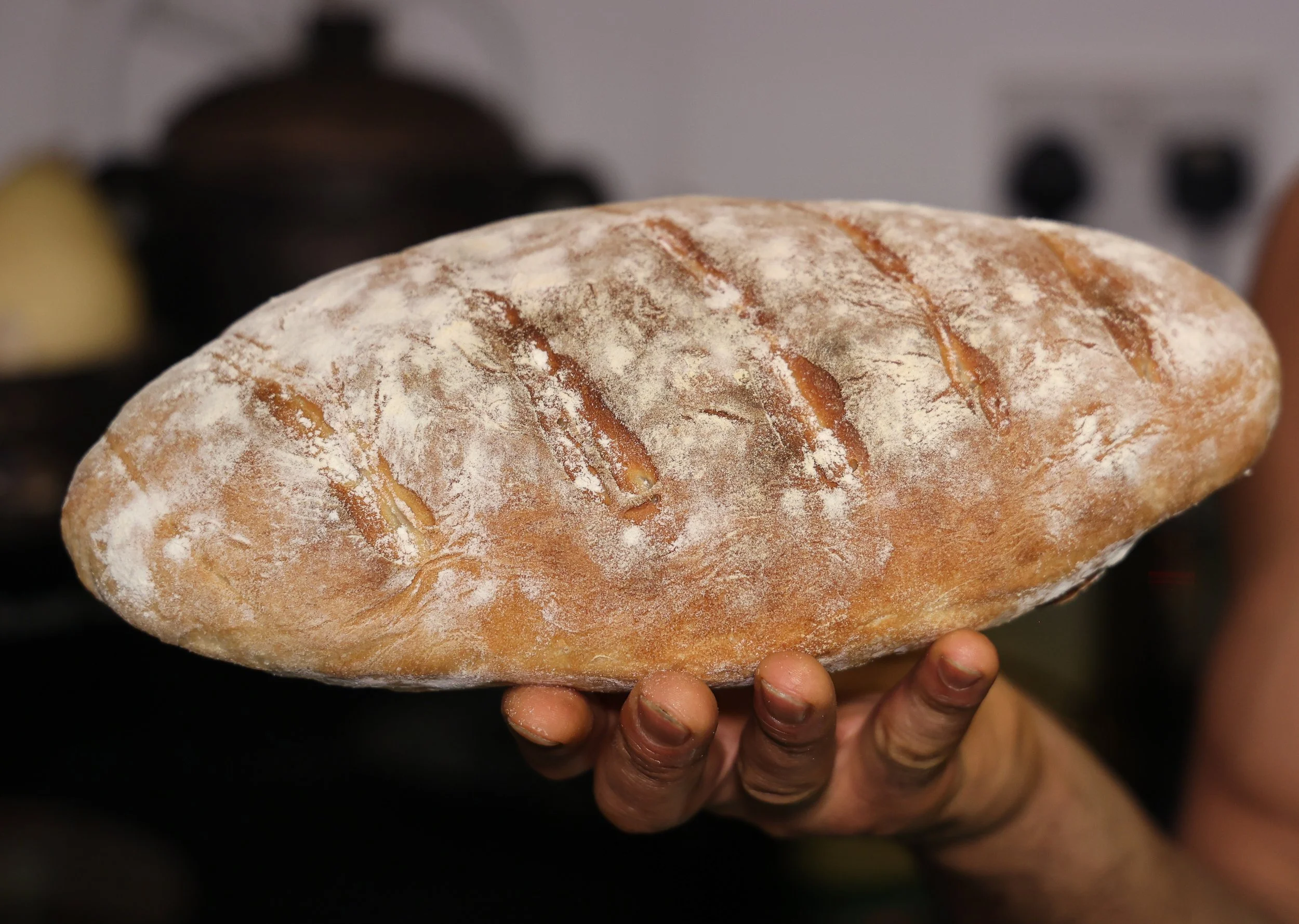 A hand holding a large loaf of rustic bread with a dusting of flour and slashes on top, in a kitchen setting.