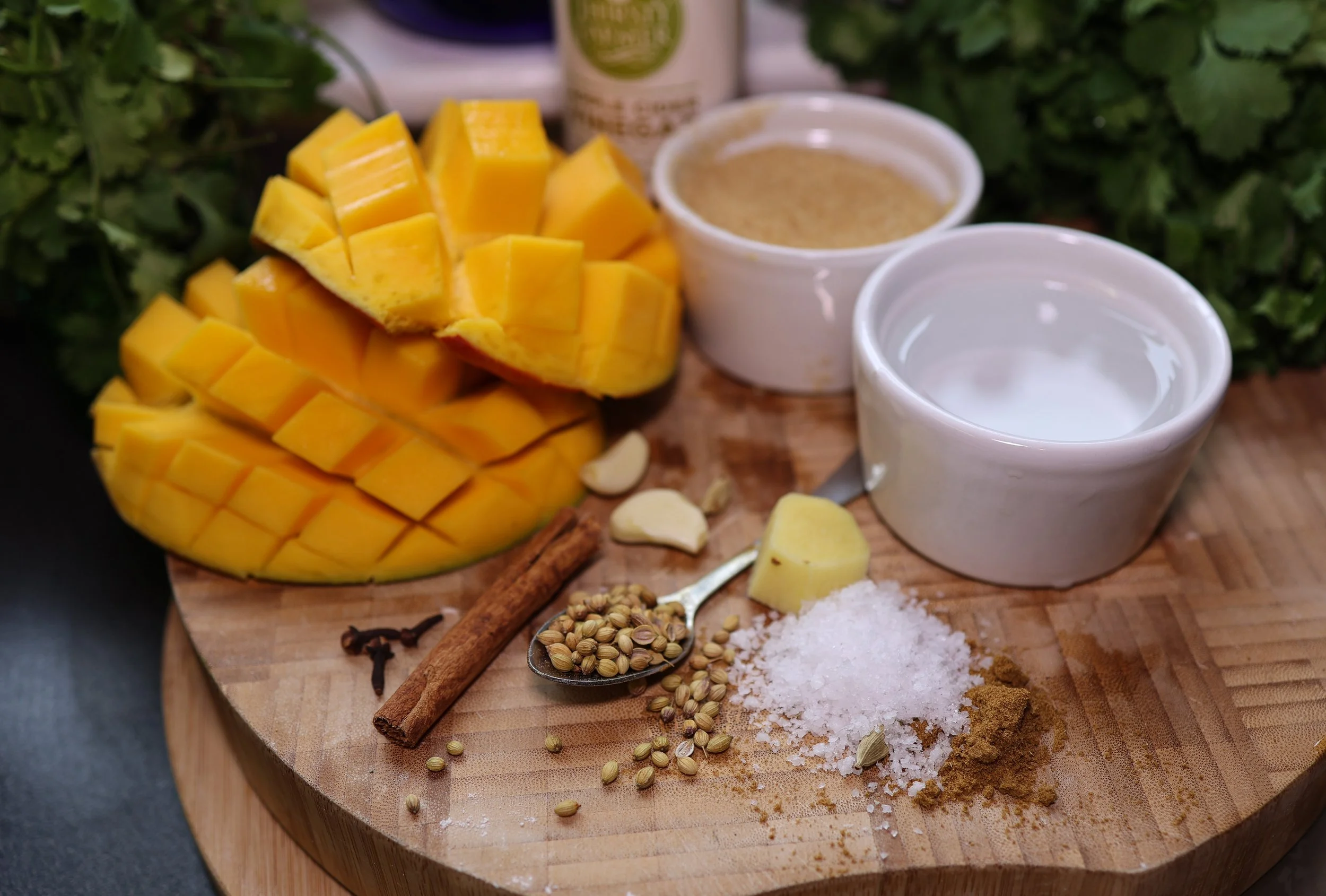 Sliced mango, garlic cloves, coriander seeds, cinnamon stick, salt, turmeric powder, ginger, coconut milk, and spices on a wooden cutting board.