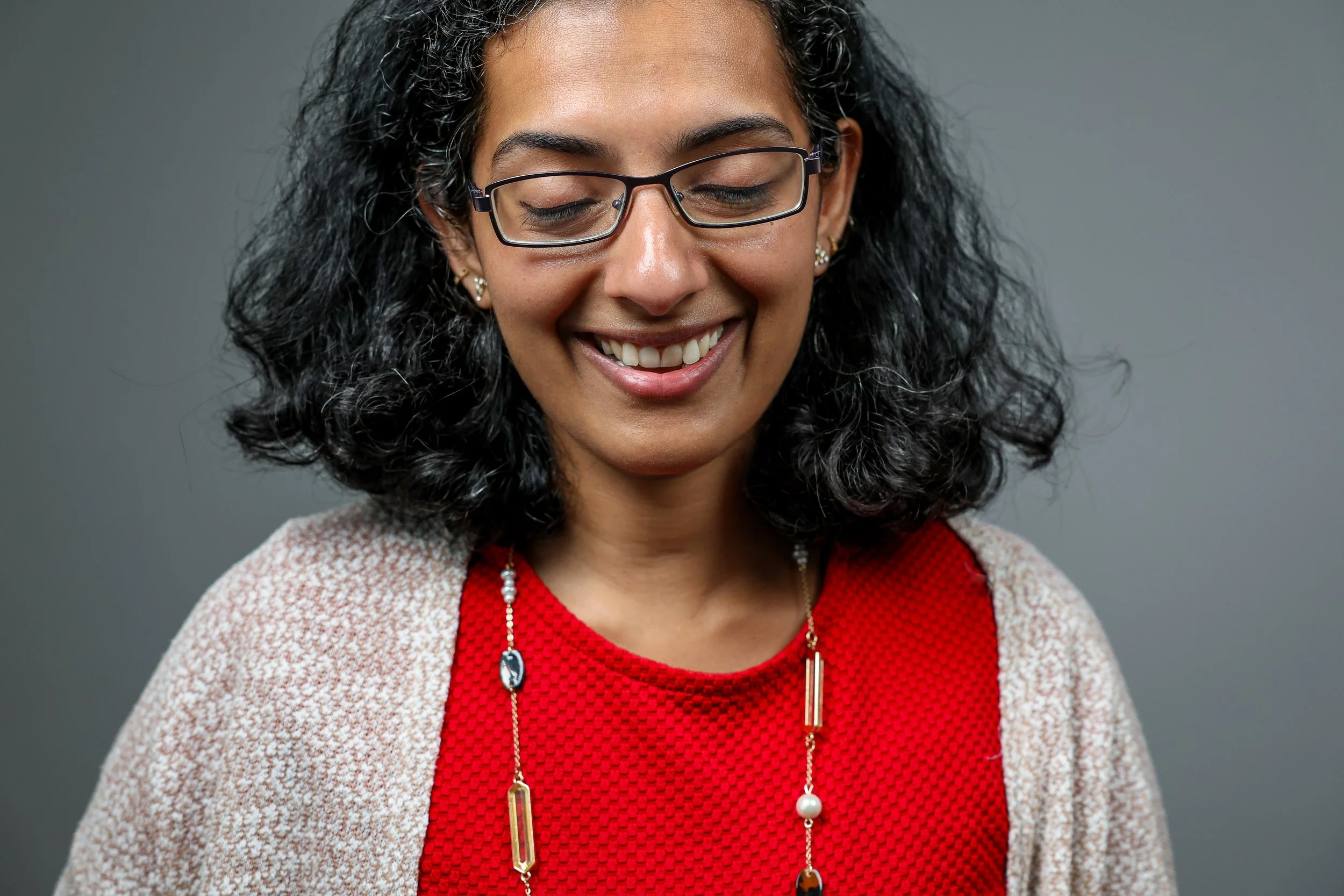 Smile of a woman with curly hair, glasses, red top, and jewelry.