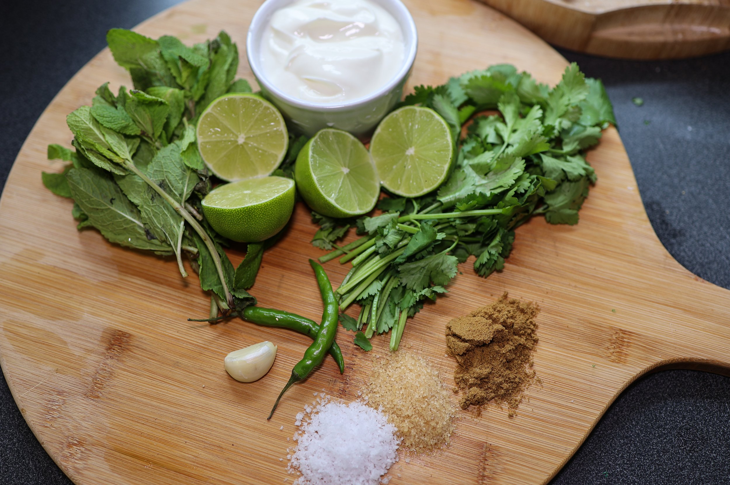Fresh herbs, limes, garlic, chilies, salt, brown sugar, and spices on a wooden cutting board for cooking.
