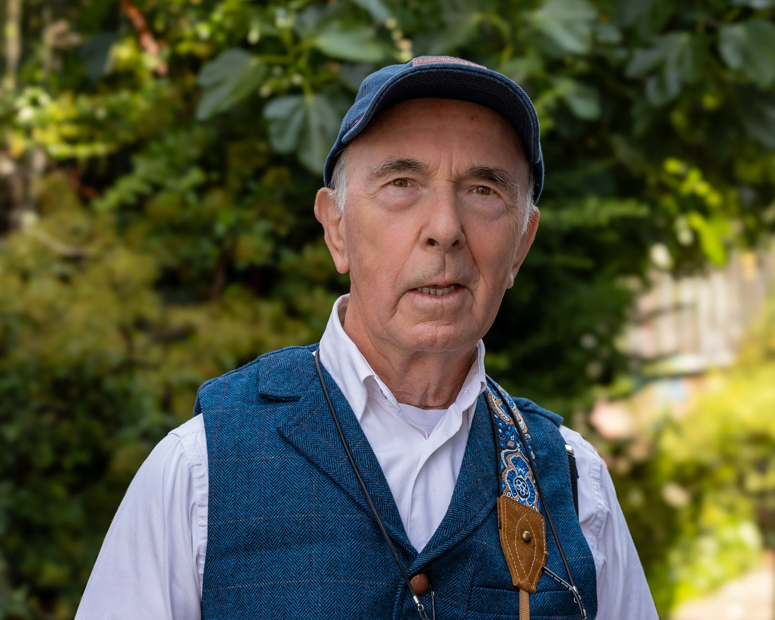 Older man wearing a navy blue cap, white shirt with a blue vest, and ornament, standing outdoors with greenery in the background.