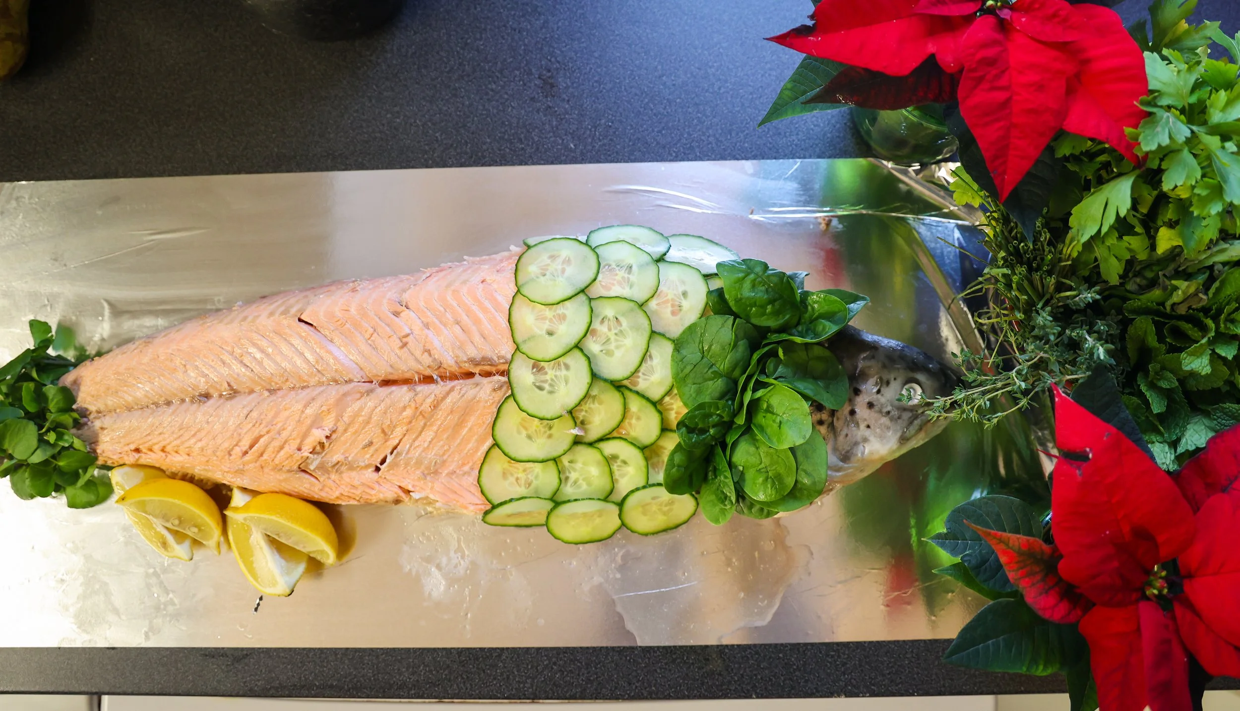 Fresh fish garnished with sliced cucumber, lemon wedges, and fresh herbs, surrounded by poinsettia and other holiday greens.
