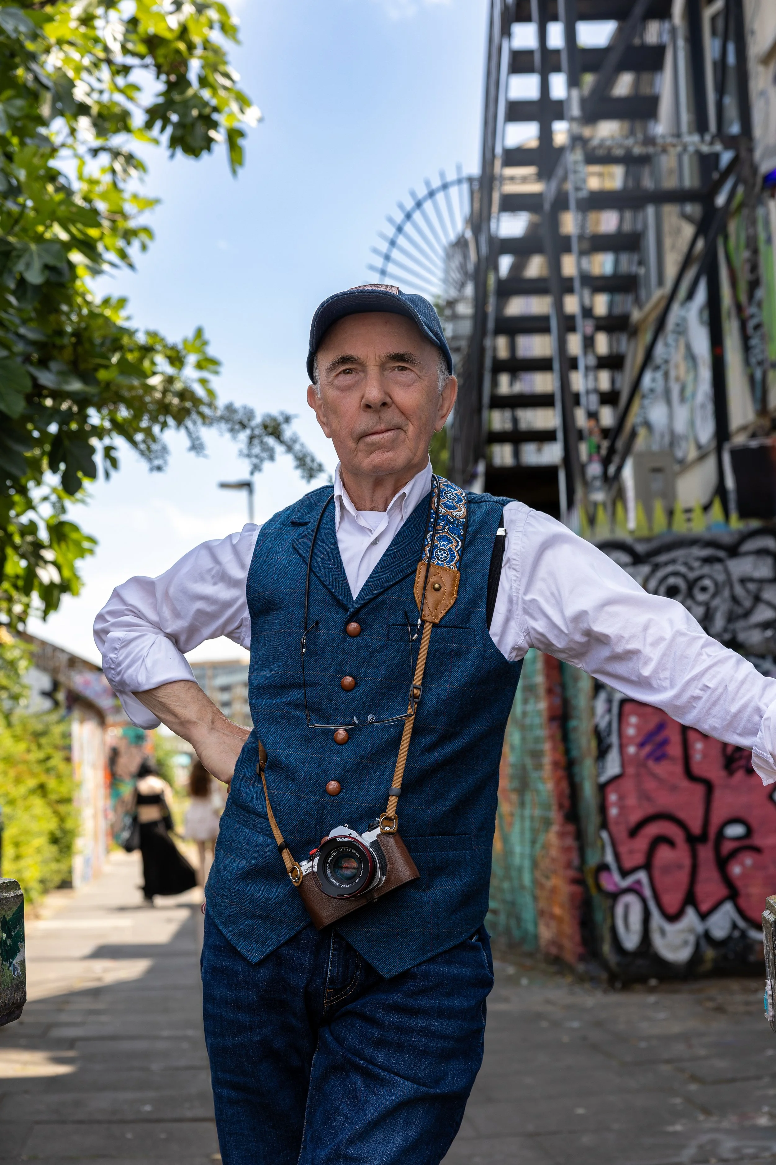 An elderly man standing confidently on a city street, wearing a white shirt, blue vest, matching pants, and a cap, with a camera hanging around his neck. Graffiti-covered walls and a staircase are visible in the background.