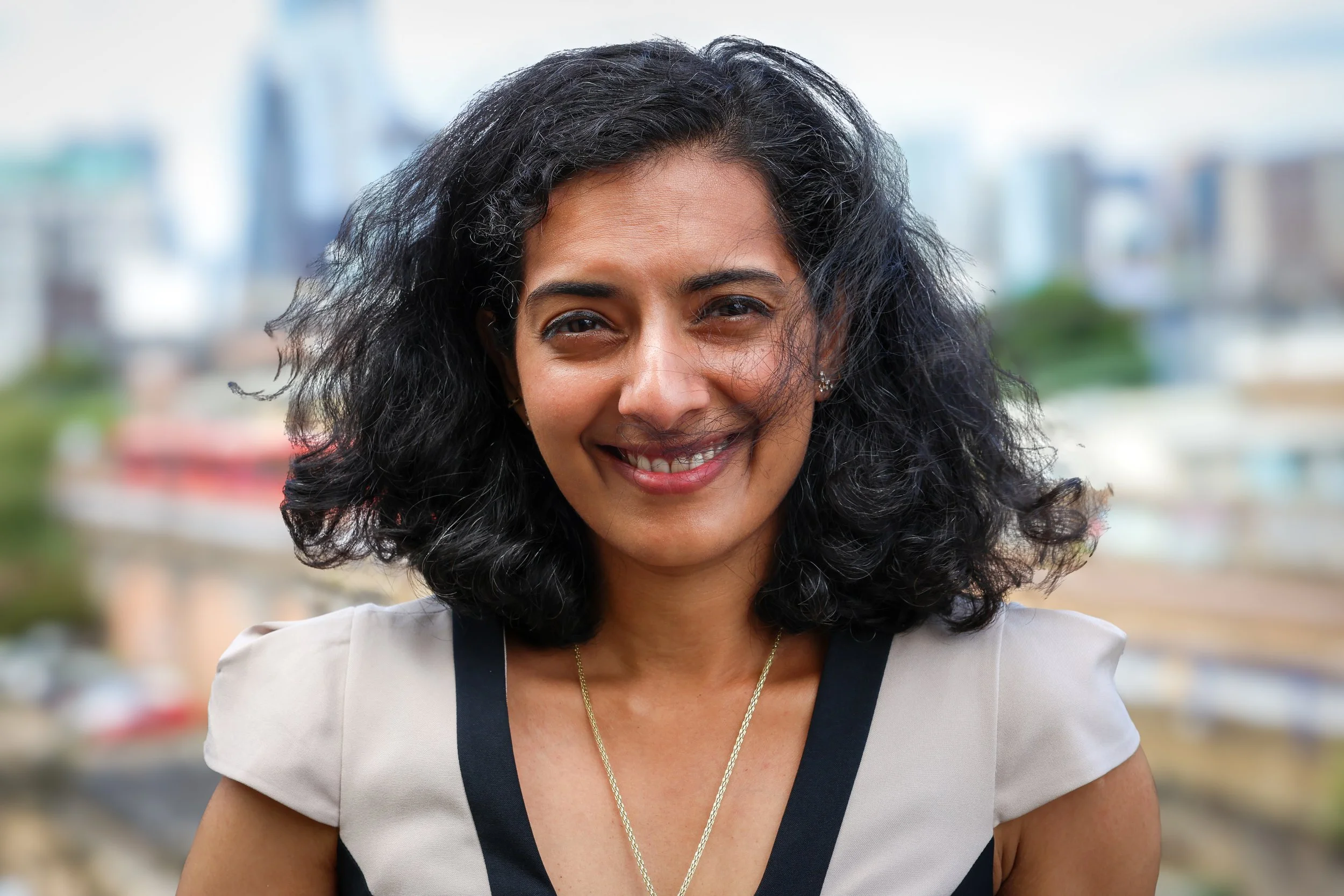 Smiling woman with curly black hair and a white and black top, standing outdoors with an urban cityscape in the background.