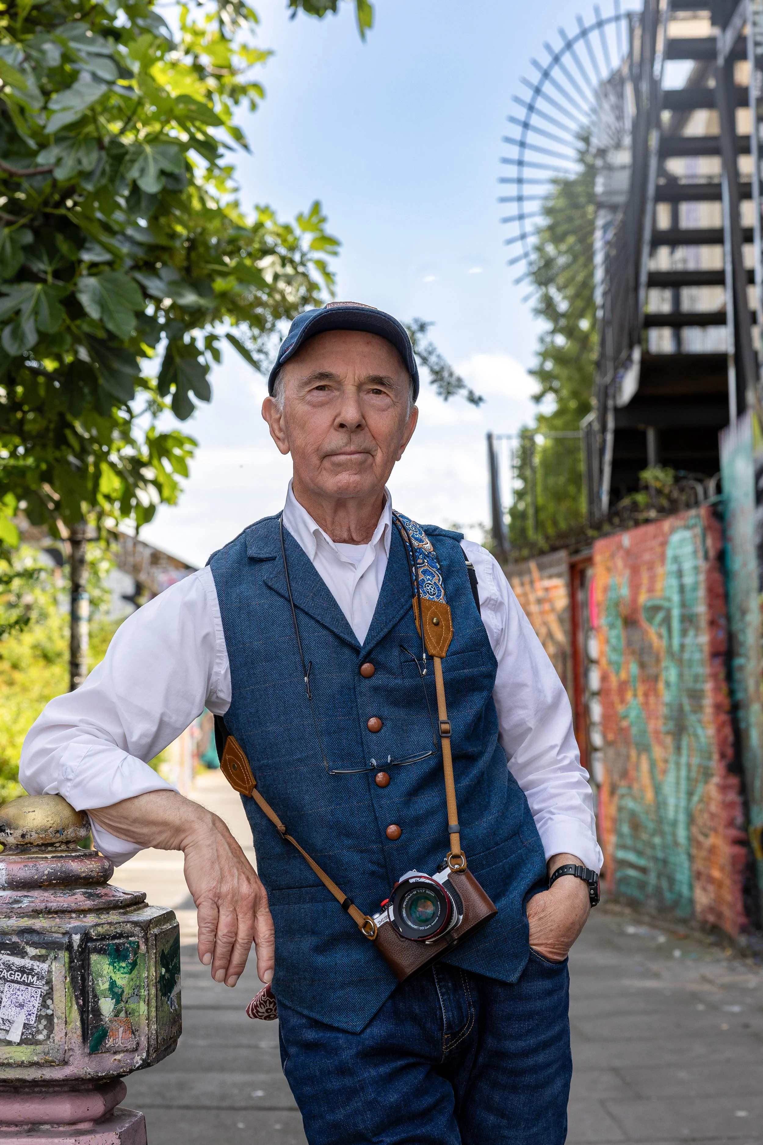 An elderly man standing outdoors near a colorful graffiti wall, wearing a white shirt, blue vest, and a camera around his neck, with a serious expression.