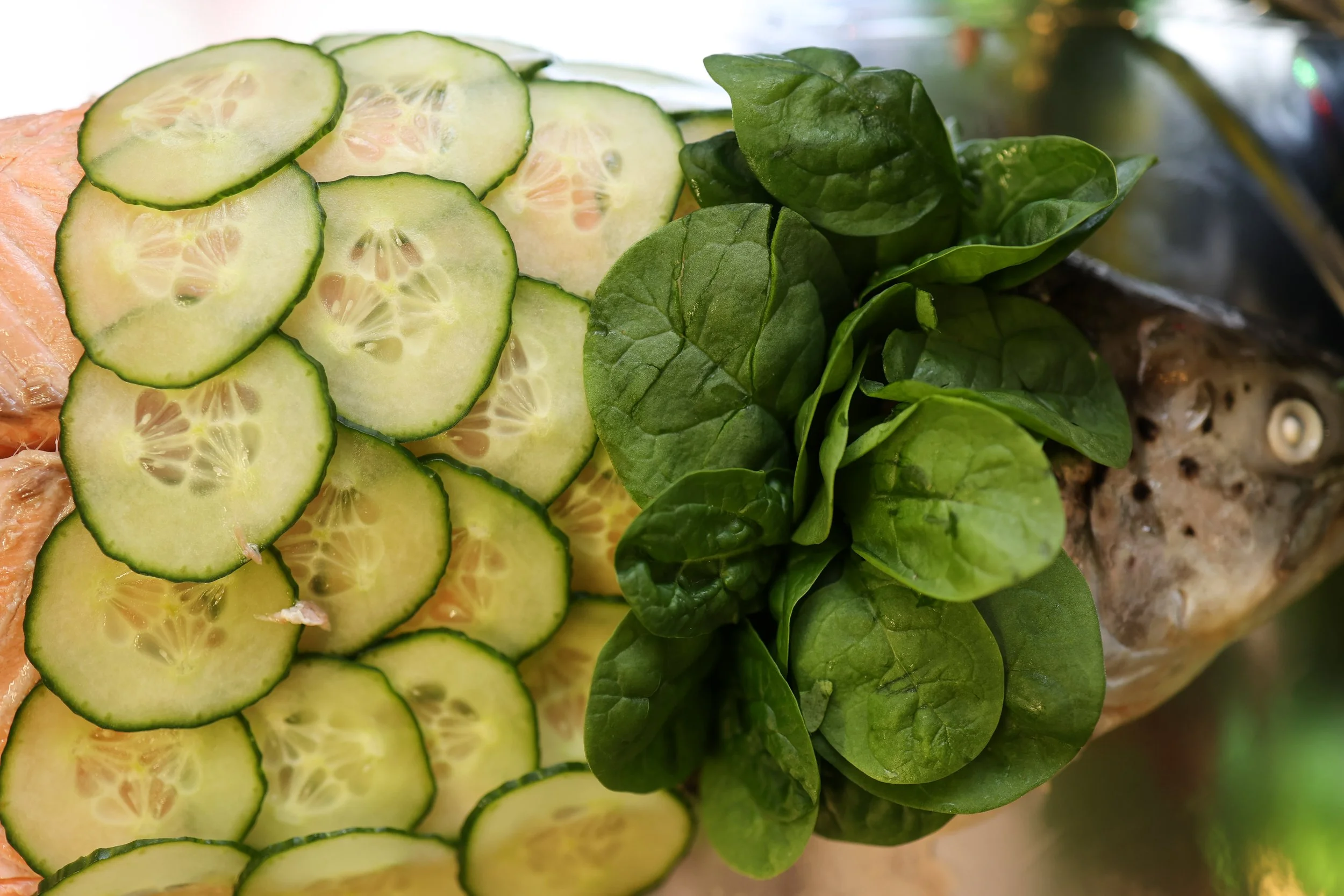 Close-up of sliced cucumber, fresh spinach leaves, and a whole fish with some lime slices on top.
