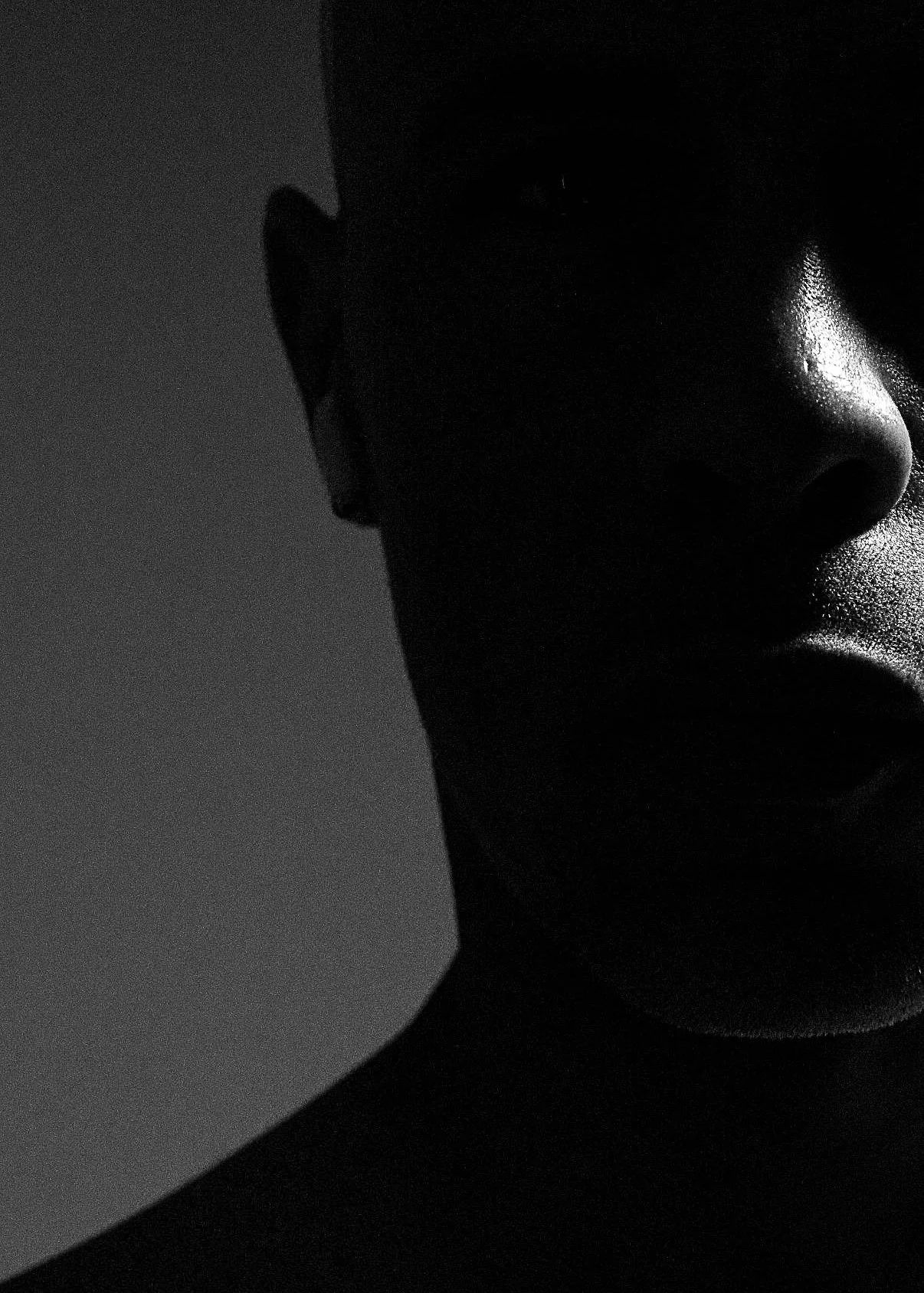 Close-up black-and-white photograph of a person's face, with prominent shadows and glossy skin, focusing on the nose and part of the lips, in soft lighting.
