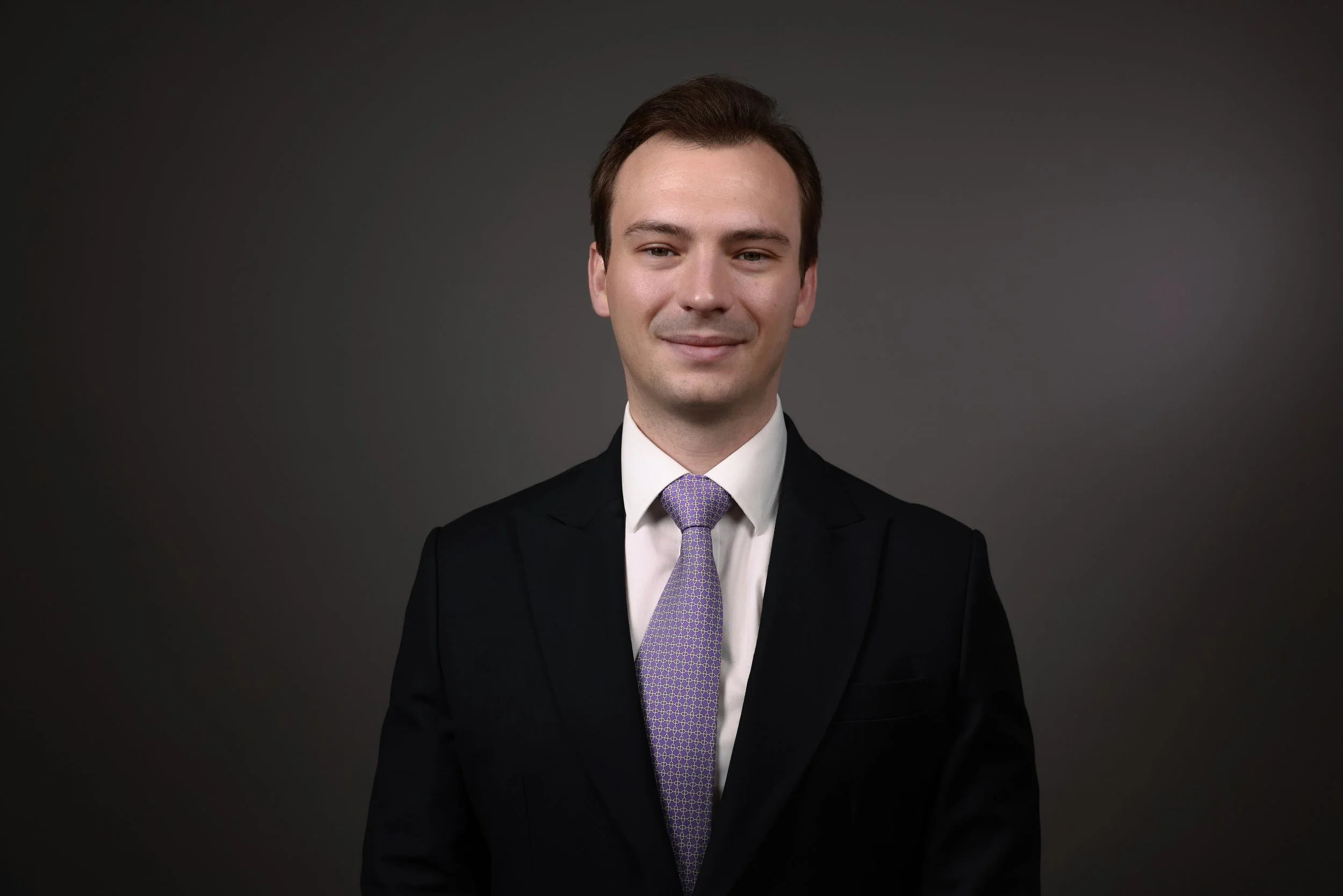 A young man in a black suit, white shirt, and patterned purple tie, standing against a dark gray background.