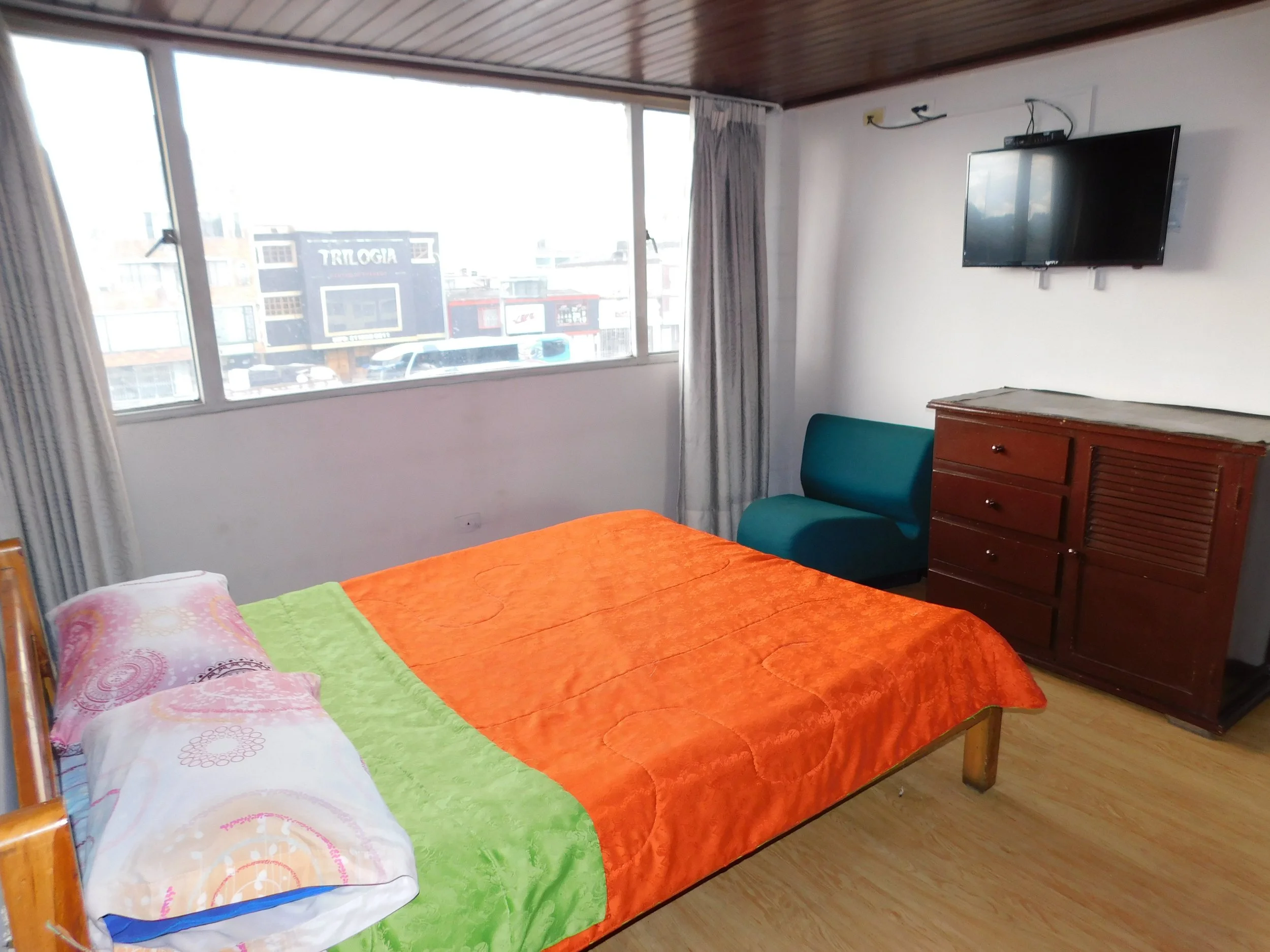 Habitación con cama, edredón naranja, almohadas, televisor en la pared, sillón azul, mueble de madera y ventana grande con cortinas.