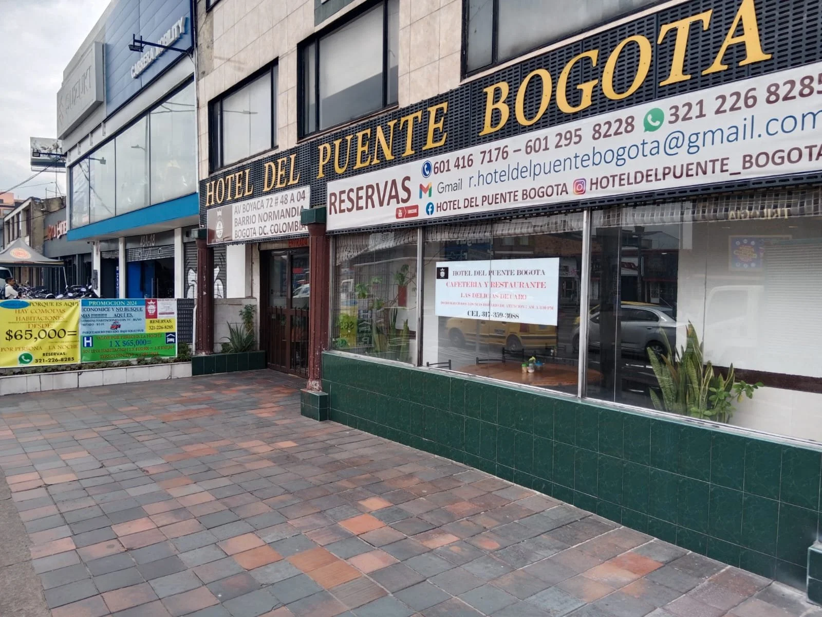 Vista exterior del Hotel del Puente Bogotá con carteles de promociones y reservaciones en la entrada, en una calle con acera de baldosas y un cielo nublado.