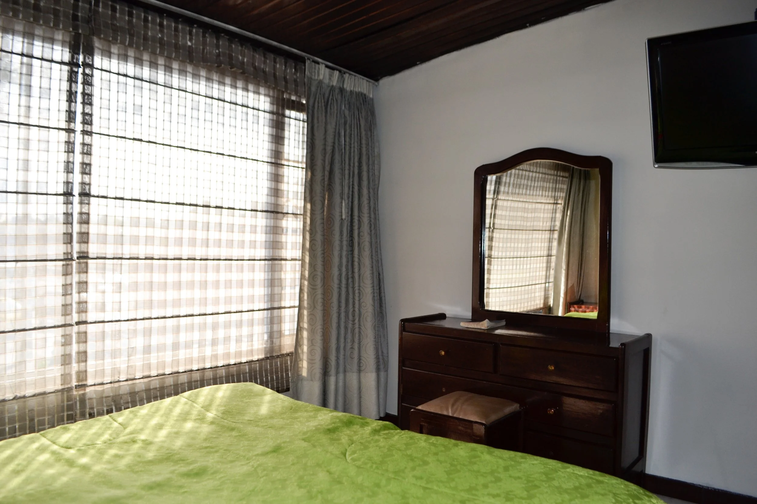 Habitación con ventana grande, cortinas y mosquiteros, ropa de cama de color verde en la cama, un mueble de madera con espejo y cajones, y una televisión en la pared.