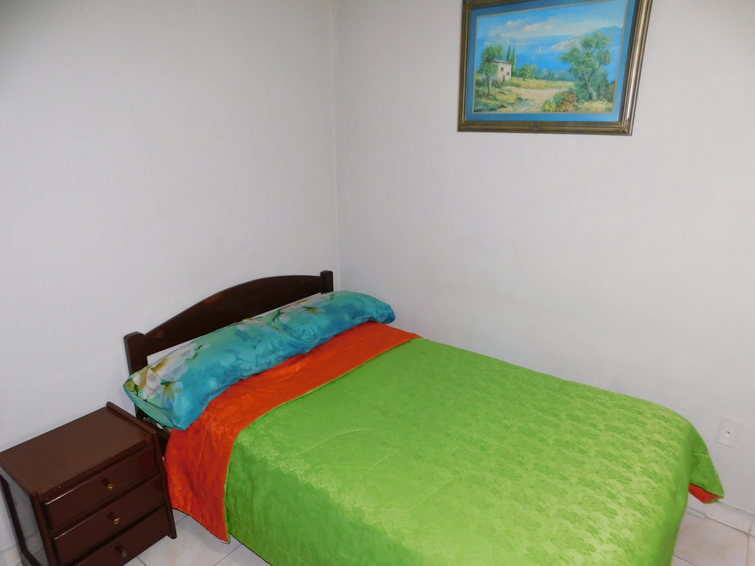 Habitación con cama de madera, sábanas y cobija de colores verde, naranja y azul, y una pintura de paisaje en la pared