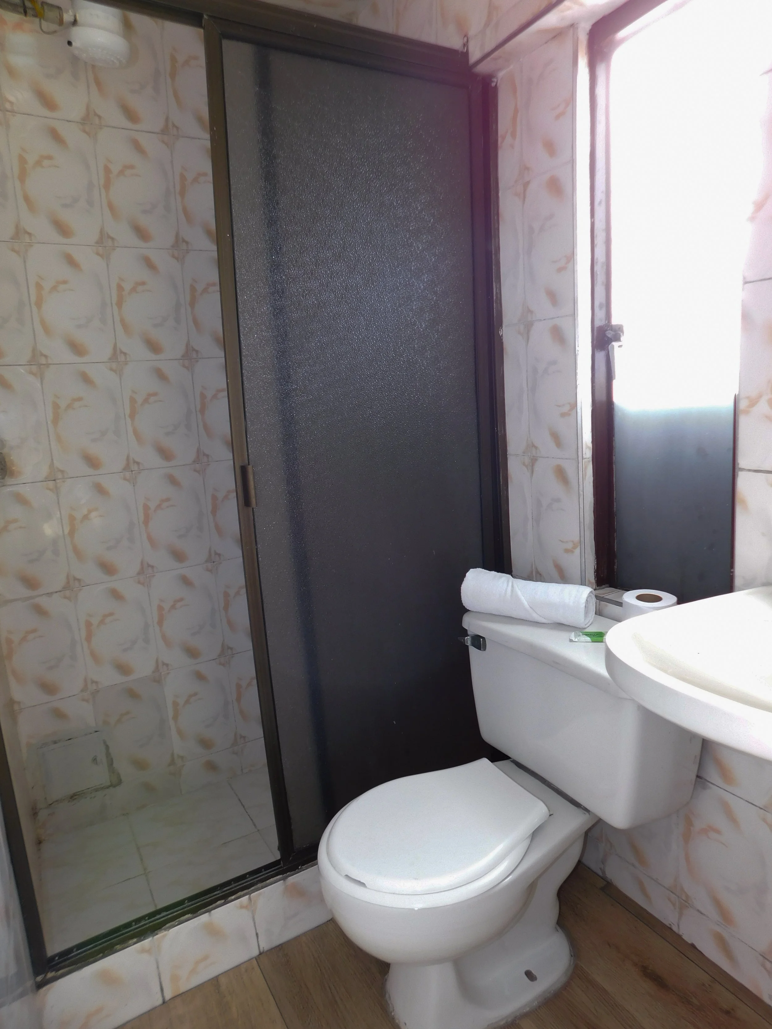 Baño con ducha, inodoro con papel y toalla encima, y lavabo, con paredes revestidas en azulejos con diseño marrón y beige, y ventana con luz natural.